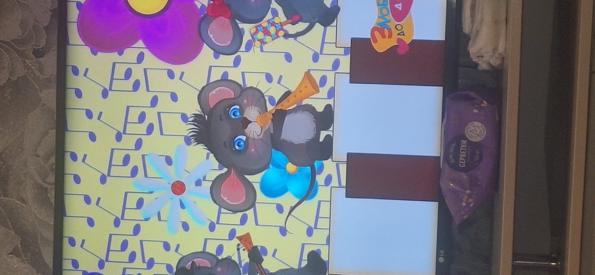 A television screen displays a cartoon of three mice playing musical instruments against a backdrop of musical notes and flowers. The leftmost mouse plays a guitar, the middle mouse plays a horn, and the rightmost mouse plays a maraca. Below the cartoon is a keyboard with white and brown keys. To the right of the keyboard is a logo with the text "З любов'ю до дітей" which translates to "With love for children". Below the screen is a purple package of wet wipes with text in Cyrillic. The television is positioned on a shelf. The background of the actual photograph is a textured wall.