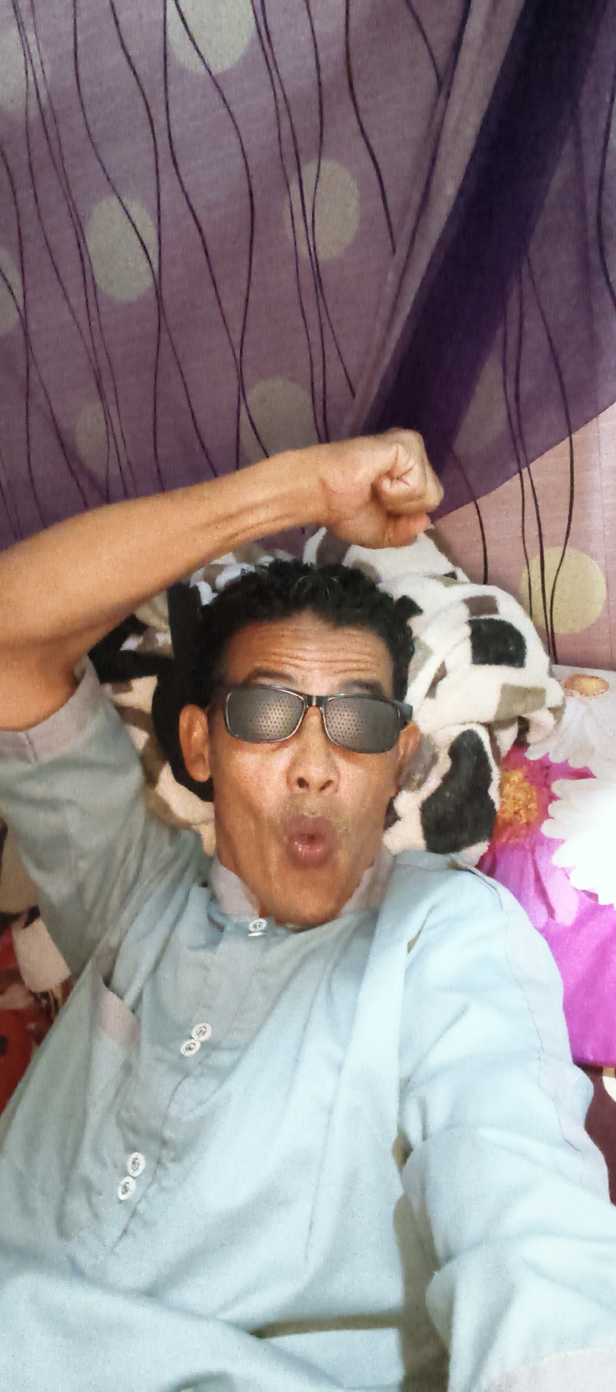 The image shows a close-up, high-angle selfie of a man with dark curly hair and glasses, wearing a light blue traditional Indonesian shirt with decorative white buttons. He is lying down on a bed or couch with a patterned blanket or fabric behind him, which features purple and cream polka dots with wavy black lines. The man is making a surprised or playful expression with his mouth open and one arm raised with a clenched fist. The lighting suggests an indoor setting. The location is specified as Muaro Jambi, Indonesia.