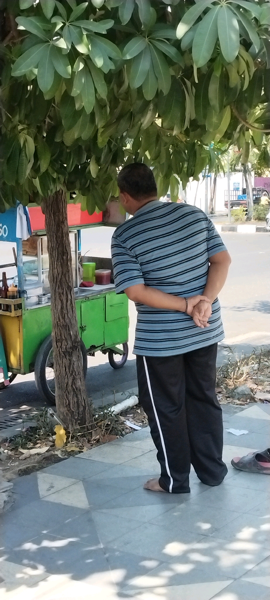 In Surabaya, Indonesia, a man stands facing away from the camera, his hands clasped behind his back, under the dappled shade of a lush tree. He is wearing a horizontally striped blue and gray t-shirt and black athletic pants with a white stripe down the side. He is barefoot. Beside him, to his left, is a green food cart with yellow accents and a red top, parked on a concrete sidewalk. Various items are visible on the cart, including green and red containers. Behind the cart, a street with vehicles and buildings can be seen. The ground is paved with light-colored tiles, and dry leaves and debris are scattered near the tree's base.