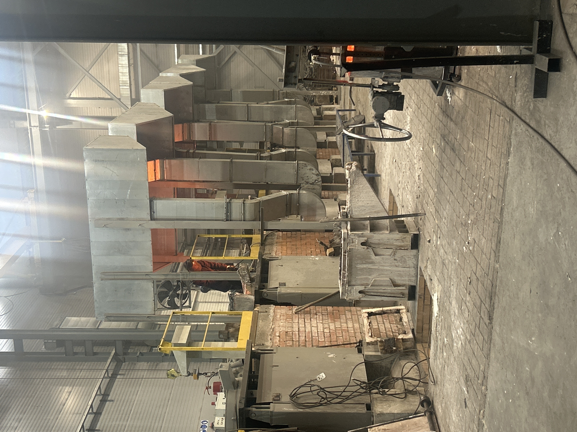 The image depicts the interior of an industrial facility, potentially a steel mill or factory, located in Noviy, Russia.
The scene showcases a long, narrow corridor with heavy machinery and ductwork extending along the ceiling and walls. The air appears hazy, possibly due to heat or industrial processes.
Large metal structures, likely furnaces or ovens, are visible along the sides of the corridor, constructed with brick and steel. The machinery exhibits signs of wear and age, with a mix of old and more modern components.
A worker is visible wearing an orange hardhat and standing near a metal platform, suggesting they are performing maintenance or repairs on the machinery.
The environment is primarily industrial and functional, with exposed pipes, wires, and support structures. The overall impression is one of a working factory involved in heavy manufacturing.