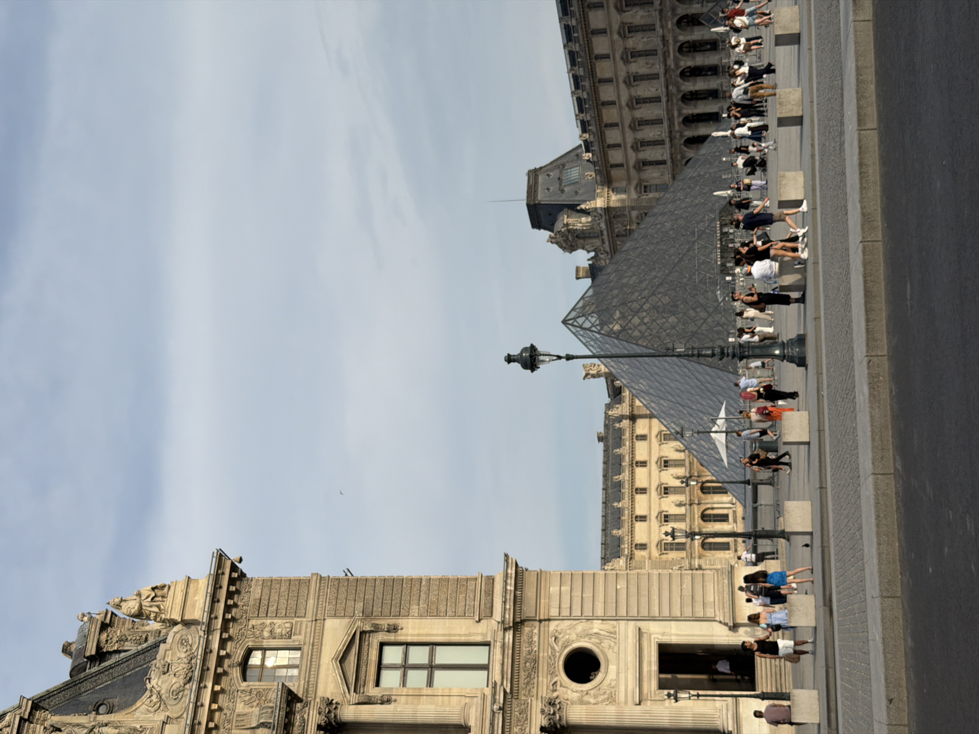 The image depicts the exterior of the Louvre Museum in Paris, France. The foreground shows a paved walkway and a street. In the middle ground, the iconic glass pyramid of the Louvre is visible, surrounded by a crowd of people. Various architectural elements of the museum, including ornate stone carvings and windows, are present on the left side of the frame. The background features more of the museum's classical architecture under a cloudy sky. People are observed walking, standing, and interacting in the plaza.