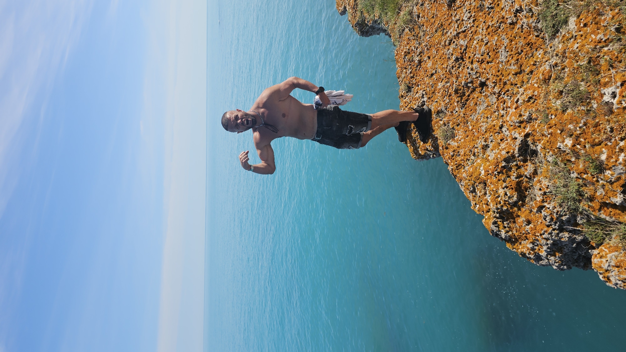 A man stands on the edge of a rocky, moss-covered cliff overlooking the turquoise sea at Kamen Bryag, Bulgaria. He is shirtless, wearing dark shorts, and flexing his bicep while shouting. He holds a white towel in his hand. The sky is clear blue.