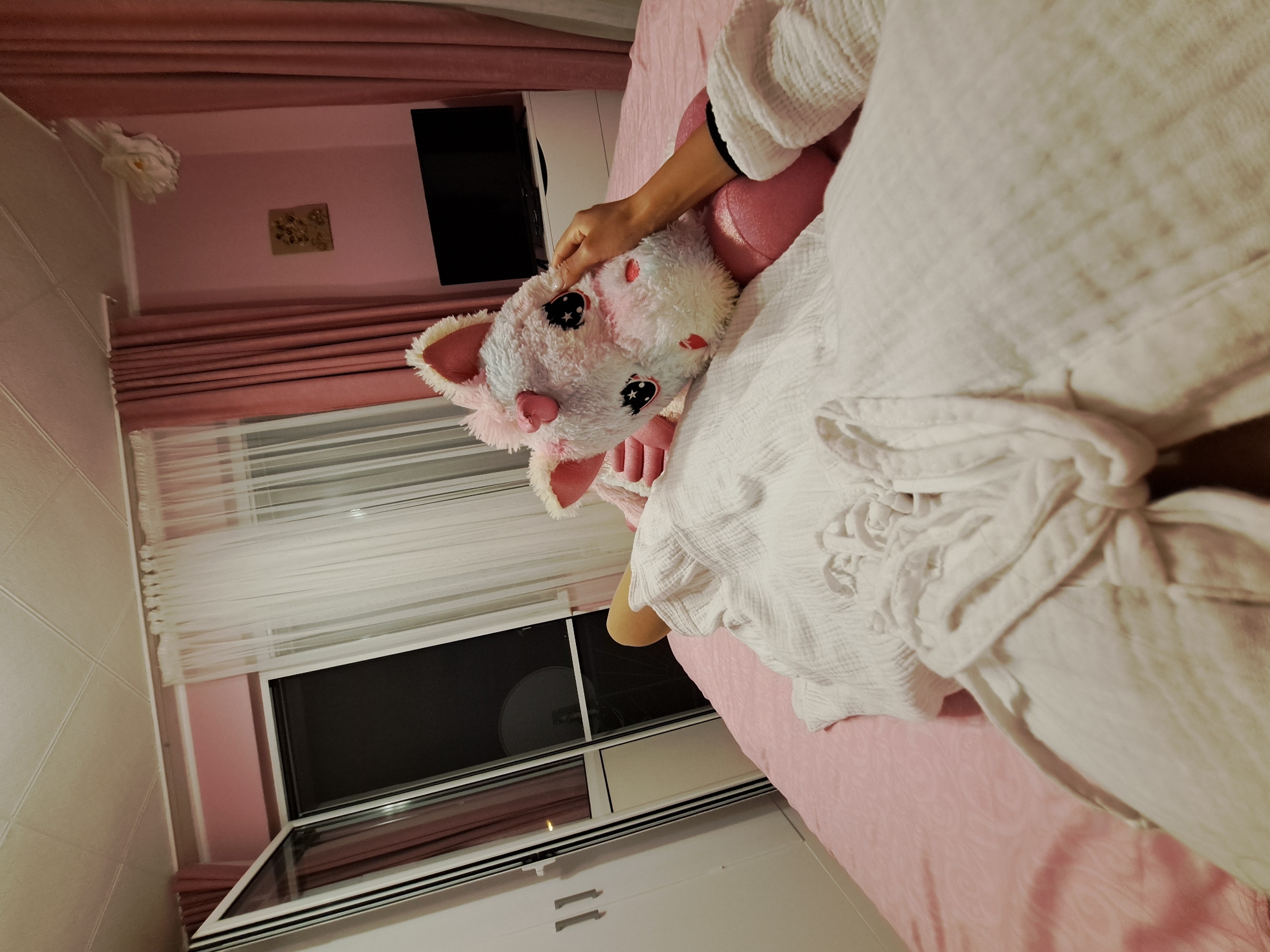 The image captures an indoor bedroom scene in the city of Unknown, Unknown, viewed from a low, first-person perspective looking down towards a person lying on a bed. The person, whose face is not visible, is partially covered by a white, textured, presumably muslin or waffle-weave, robe with a visible tied waist. One bare leg, bent at the knee, emerges from beneath the robe. The person's right arm extends forward, with the hand gently touching the face of a large, plush unicorn.
The unicorn is multi-colored, predominantly white, light pink, and light blue, with large black, sparkly eyes featuring white starbursts, a pink horn, and pink inner ears. It rests on the pink bedding, which features a subtle patterned texture.
The room's walls are painted a light pink. To the left, a white two-door wardrobe with silver horizontal handles is visible. Adjacent to the wardrobe is a white-framed glass balcony door, which is partially ajar, revealing a dark exterior with a faintly visible satellite dish in the distance. Sheer white curtains hang behind heavy pink drapes, which frame the balcony door.
On the far wall, a black flat-screen television is mounted above a white low-profile dresser with at least two white drawers. Above the television, a small, square piece of textured artwork, possibly depicting floral elements in relief, is affixed to the wall. In the upper right corner of the room, near the ceiling, a large white decorative floral element, appearing to be made of fabric or paper, is mounted. The ceiling is white with a grid-like paneling pattern. The room is illuminated by warm interior lighting, indicating an evening or nighttime setting.