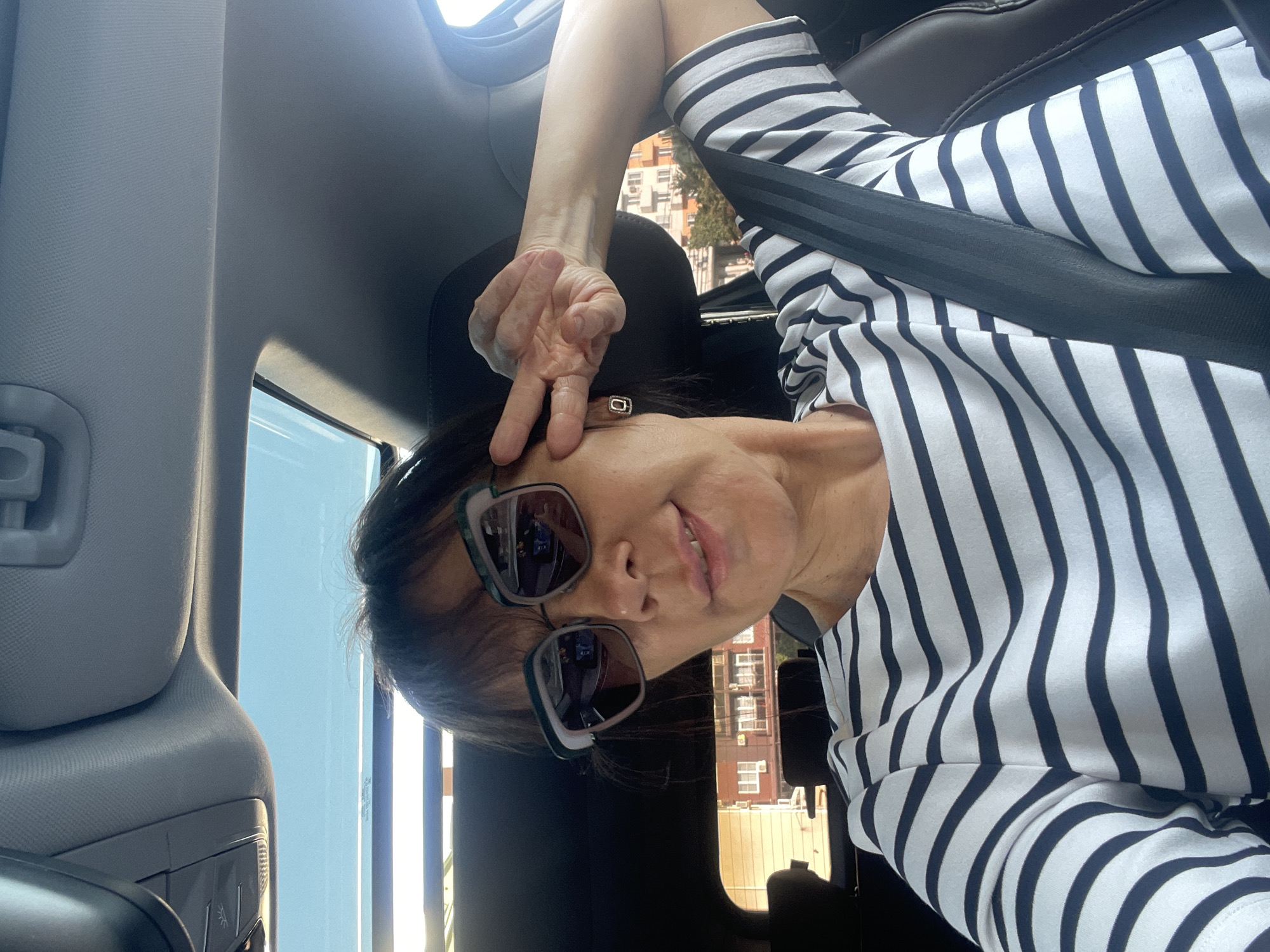 The image is a selfie taken from inside a car, featuring a woman with sunglasses on. She is wearing a black and white striped long-sleeved shirt. Her right arm is raised, with her fingers forming a peace sign. The background shows buildings, suggesting an urban or suburban setting. The sun appears to be shining, as indicated by the bright lighting and shadows. The interior of the car is visible, including a seatbelt and part of the dashboard. The orientation of the photo is rotated.
