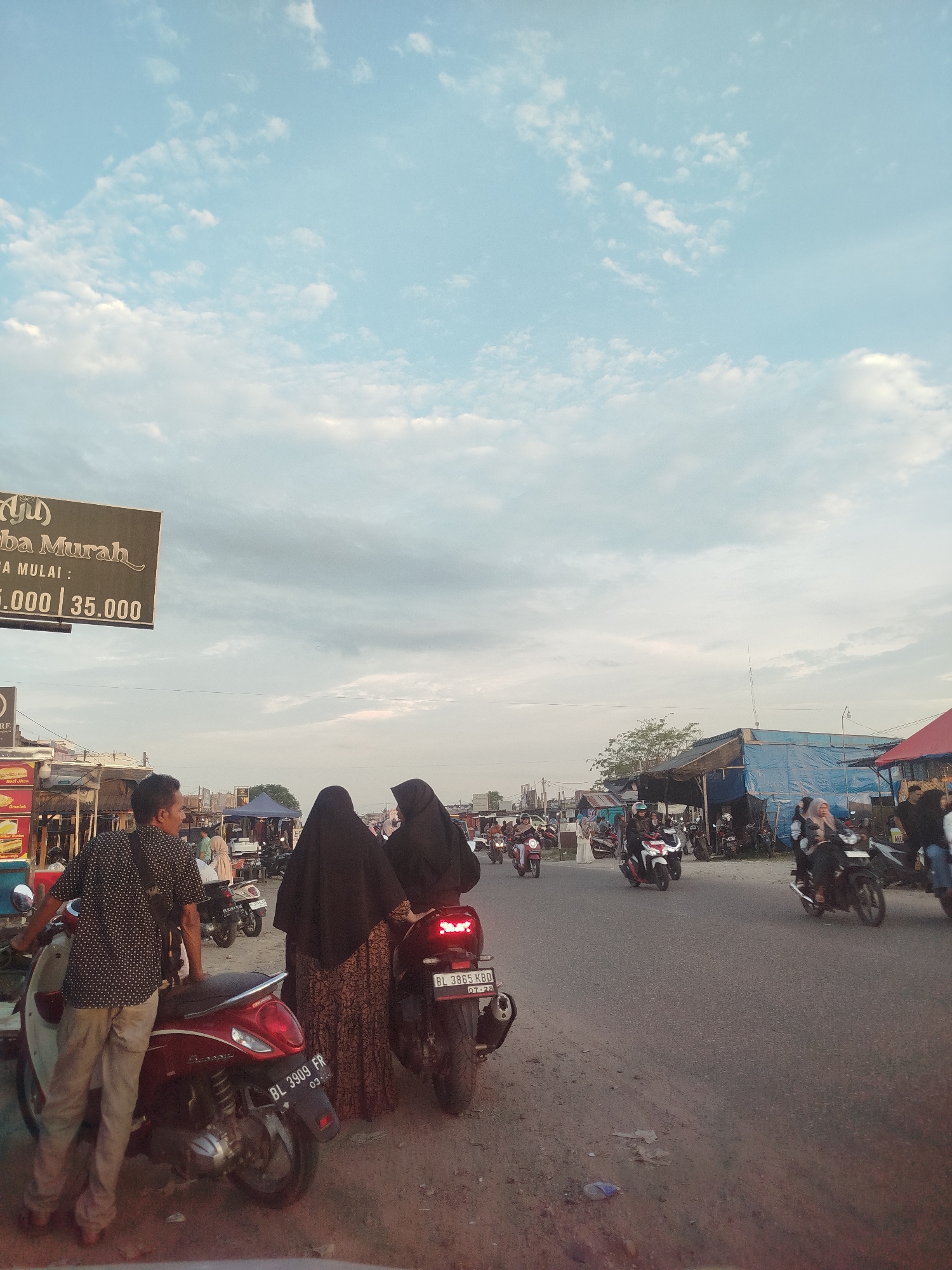 The image depicts a street scene in Peurupok, Indonesia. Several people are present, including two women wearing hijabs and long dresses standing near parked motorbikes on the side of the road. A man stands by another motorbike, looking toward the road. Motorbikes are being driven on the road. There are shops and stalls along the roadside. A sign is visible with the words "Murah" and a price range. The sky is partly cloudy.