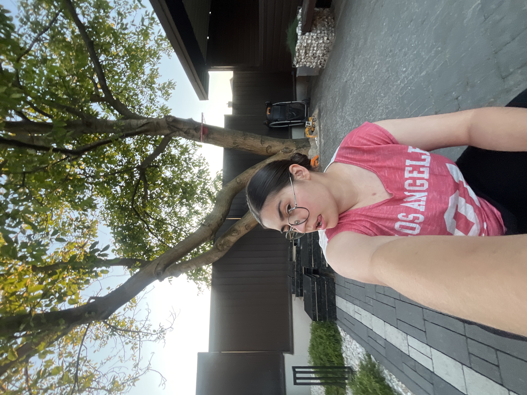 A young woman with dark hair and glasses is taking a selfie outdoors. She is wearing a pink t-shirt with white text that reads "LOS ANGELES". The setting appears to be a backyard or patio area with a modern house. A large tree with green leaves is visible on the left side of the frame, and a paved area with a geometric pattern is in the foreground. In the background, there is a dark-colored building with wooden siding and some decorative stone elements. A child's toy bulldozer and a black stroller are also visible in the background. The lighting suggests it is late afternoon or early evening, with the sun casting a warm glow. The woman's expression is relaxed and casual. The location context is provided as Koroviia, Ukraine.