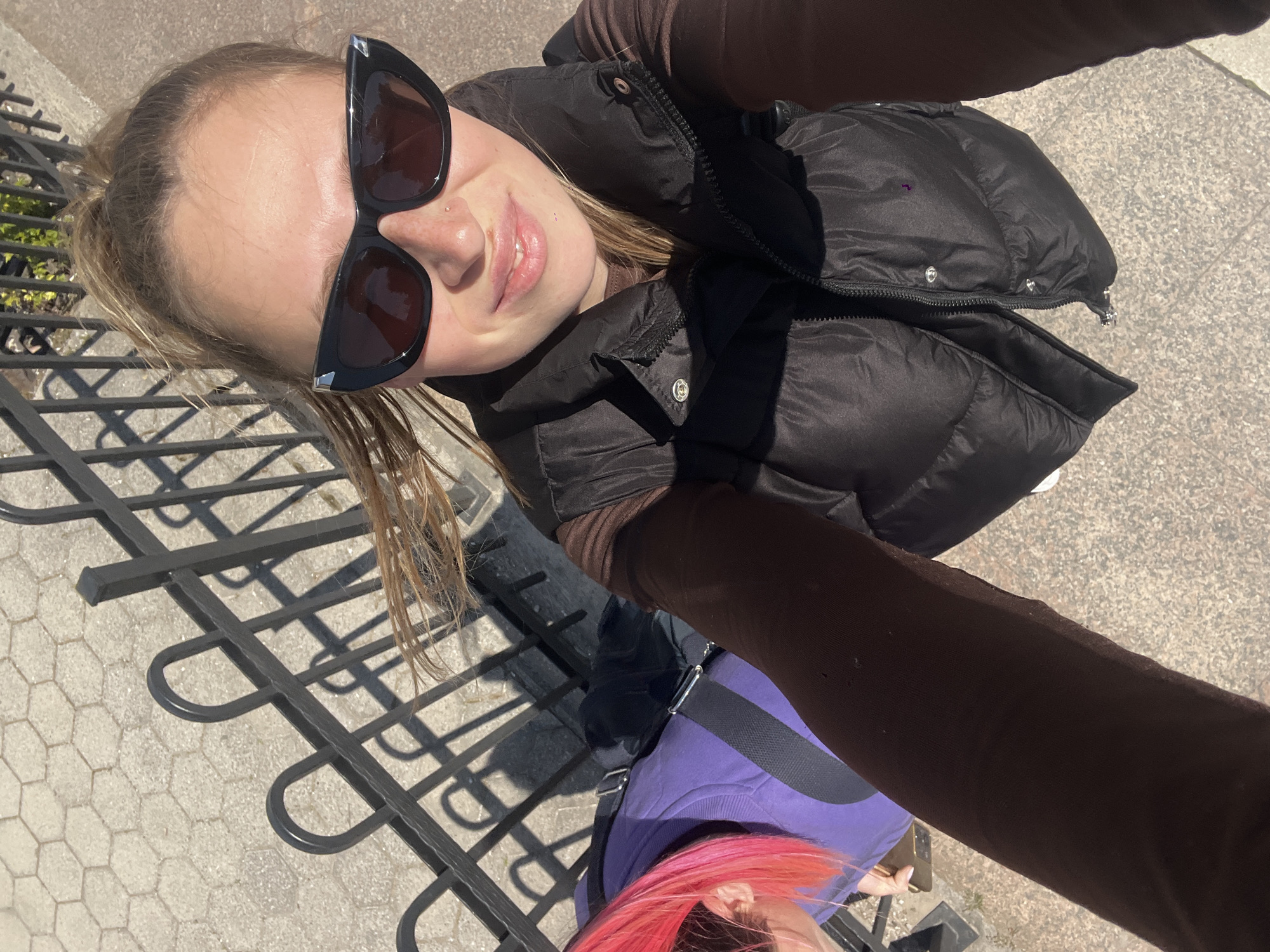A selfie shows two people standing near a metal fence on a paved surface. The primary subject is fair-skinned, wearing black sunglasses and a black puffer jacket over a brown long-sleeved shirt. The person is smiling slightly at the camera. The secondary subject is visible from the chest down. They are wearing a purple shirt with a black strap across the torso. They have pink hair. The angle of the photograph suggests that the primary subject is holding the camera. The presence of a metal fence and paving stones suggests an outdoor location, possibly a park or pedestrian area. The precise location within Budva, Montenegro, cannot be determined from this single image.