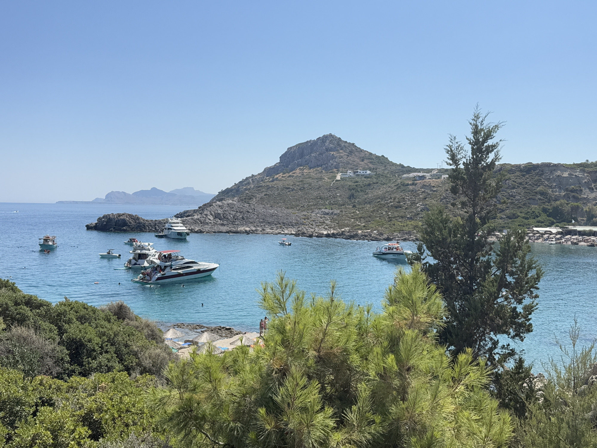 This is a photograph of a bay in the city of Rhodes, Greece. The bay is filled with clear blue water and several boats, including speedboats and yachts. Some of these boats have people on them, some of whom appear to be swimming or enjoying the water. The shoreline is lined with trees and vegetation, and a small beach with umbrellas is visible in the distance. In the background, a large, rocky hill dominates the landscape, with some buildings visible on its slopes. The sky is clear and blue, indicating a sunny day.