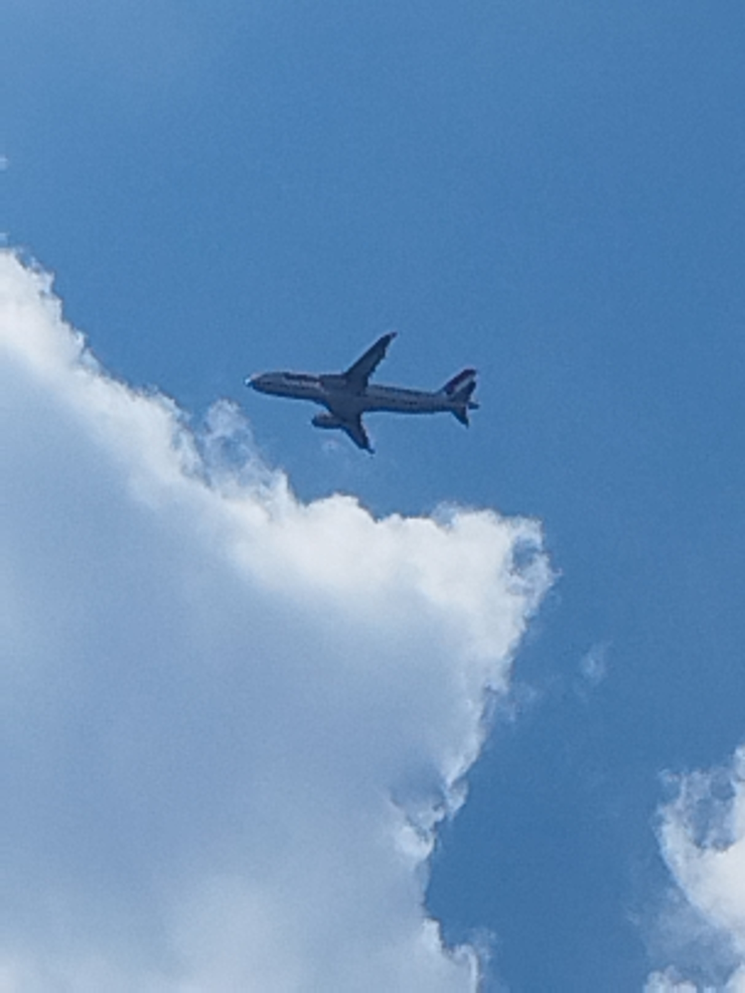 A passenger airplane is observed flying against a clear blue sky with some white clouds. The airplane is silver with red accents on its tail fins. It is positioned in the upper right quadrant of the image, viewed from below and slightly from the side, indicating it is ascending. The image appears to have been taken on a sunny day. The location is the city of Buftea, Romania.
