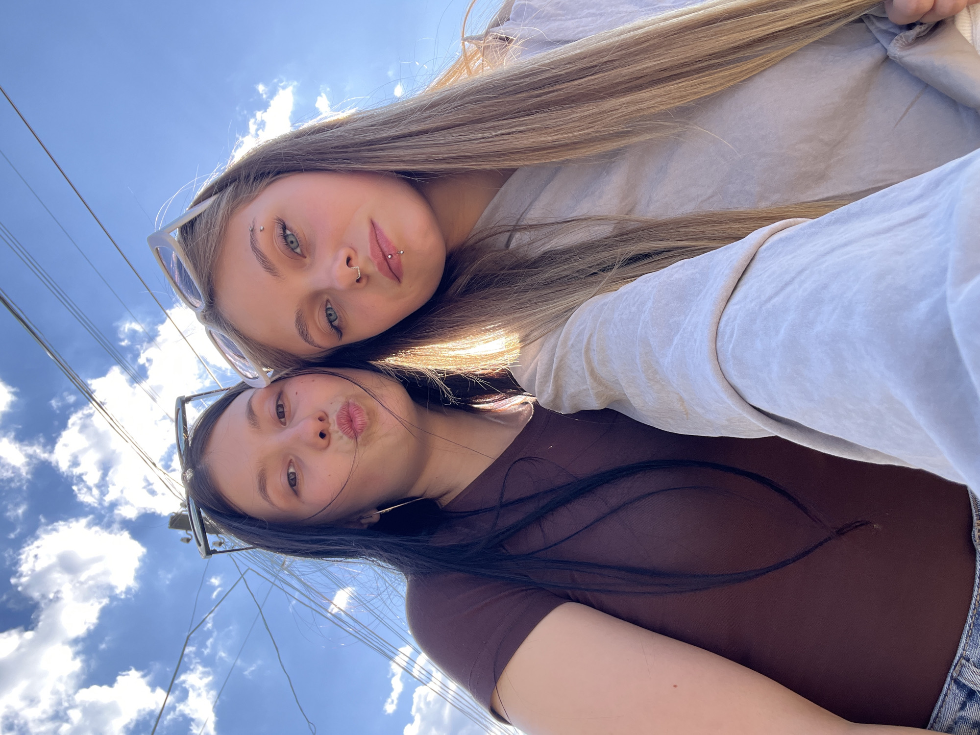 Two young women are posing for a selfie outdoors, likely on a bright day. The woman on top has long, straight blonde hair, fair skin, and blue eyes. She has piercings on her eyebrow, nose, and lip. She is wearing sunglasses perched on her head and a light-colored, long-sleeved shirt. The woman below has long, dark hair, fair skin, and brown eyes. She has sunglasses perched on her head and is pouting her lips for the photo. She is wearing a brown t-shirt. The background shows a clear, blue sky with scattered white clouds, along with power lines and utility poles. The location is likely Фоки, Russia.