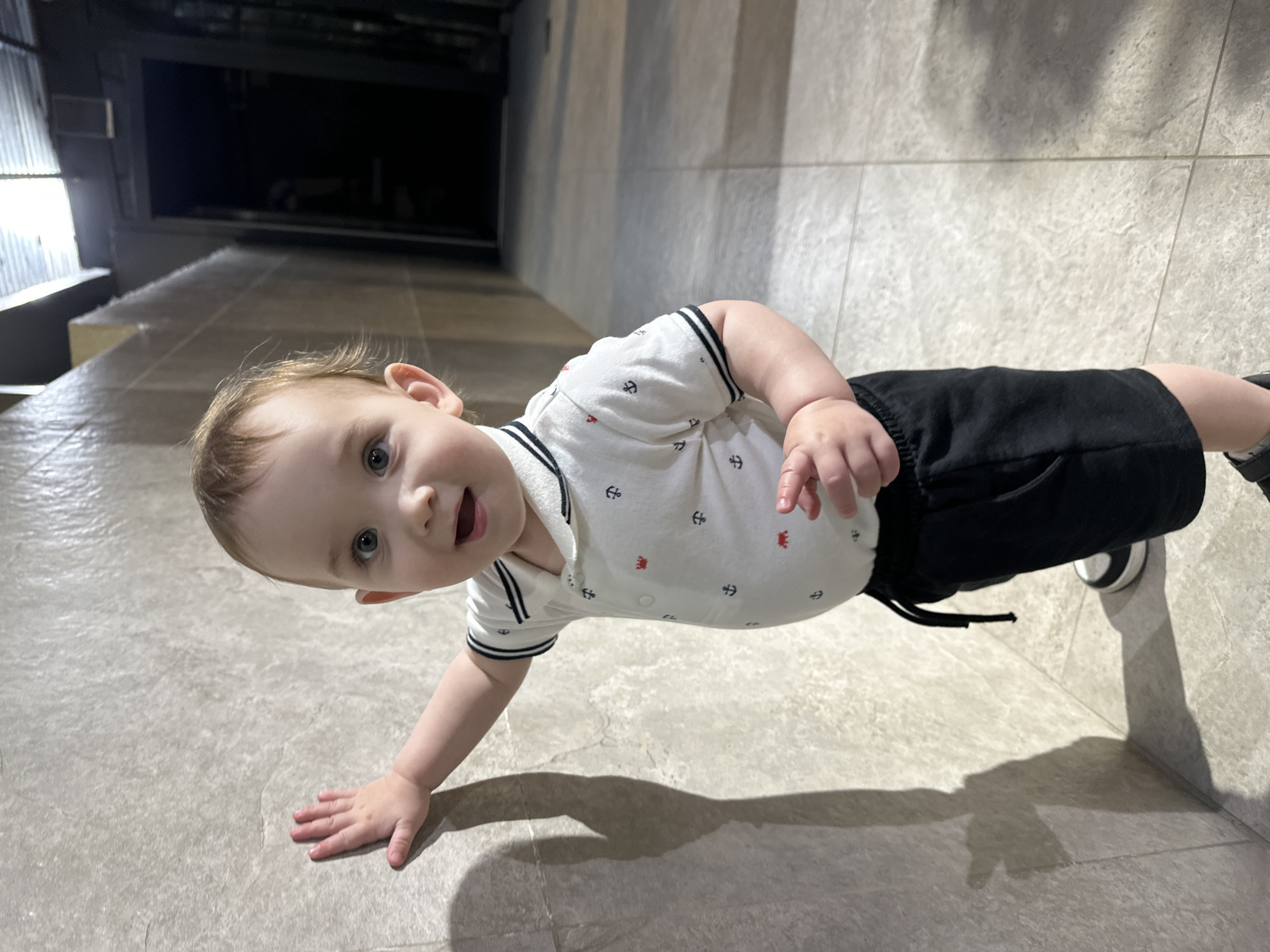 A young child, likely a toddler, stands next to a light-colored tiled wall in an indoor setting. The child is wearing a white polo shirt with a pattern of small red crabs and black anchors, along with black shorts and sneakers. The child's left hand is touching the wall, casting a shadow. The background shows a hallway with tiled flooring and dark doorways or passages. The lighting appears to be artificial, creating distinct shadows. The exact location within Chișinău, Moldova cannot be determined from the image alone.