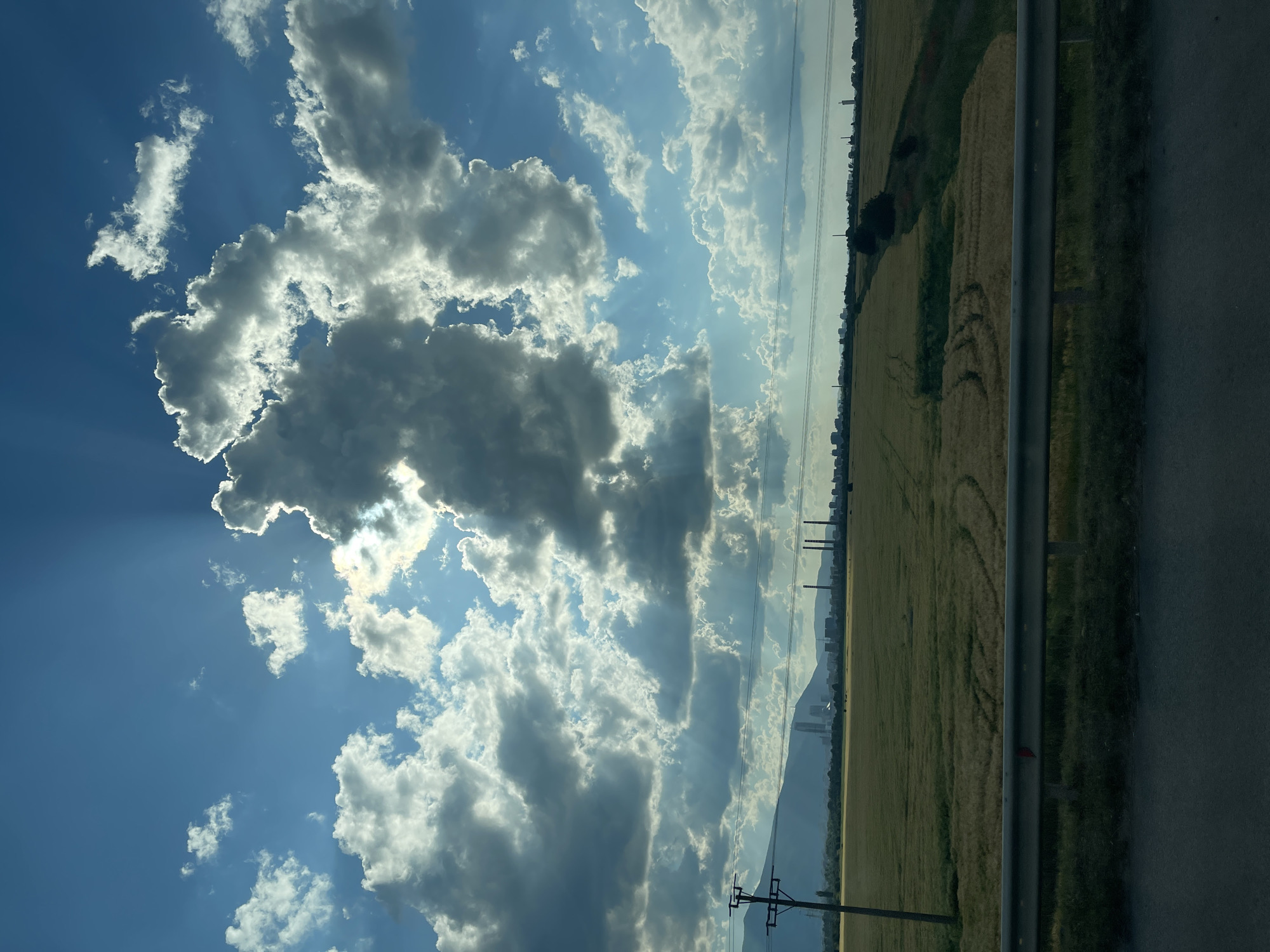 A wide-angle landscape photograph captures a rural scene with a distant industrial and urban complex under a dramatic sky. The upper two-thirds of the image are dominated by a vibrant blue sky filled with large, volumetric cumulus clouds. Sunlight breaks through the clouds from the right side of the frame, creating visible crepuscular rays that fan across the sky. The edges of the clouds are brightly illuminated, contrasting with their shadowed undersides.
In the midground, a vast field of golden-yellow crops, likely wheat, extends horizontally. Faint, curved patterns suggestive of vehicle tracks or agricultural activity are visible within the field in the lower-right quadrant. A wooden utility pole with multiple crossarms and electrical wires stands prominently on the left side of the midground. A narrow, darker strip of vegetation or a ditch runs through the field from the center-right towards the distant horizon.
The foreground features a silver metal guardrail running horizontally across the bottom third of the image, positioned above a dark asphalt road surface, suggesting the photograph was taken from a vehicle.
In the distant background, a city skyline identifiable as the area of Kazichene, Bulgaria, is visible. This includes a cluster of taller, modern-looking buildings on the left, and further to the right, a prominent industrial complex featuring three large, distinct chimneys or smokestacks, indicative of a power plant or heavy industry. Additional smaller industrial structures and general urban outlines are discernible along the horizon. A range of mountains forms the furthest backdrop, appearing as dark, hazy silhouettes beneath the sky. There are no people visible in the scene.