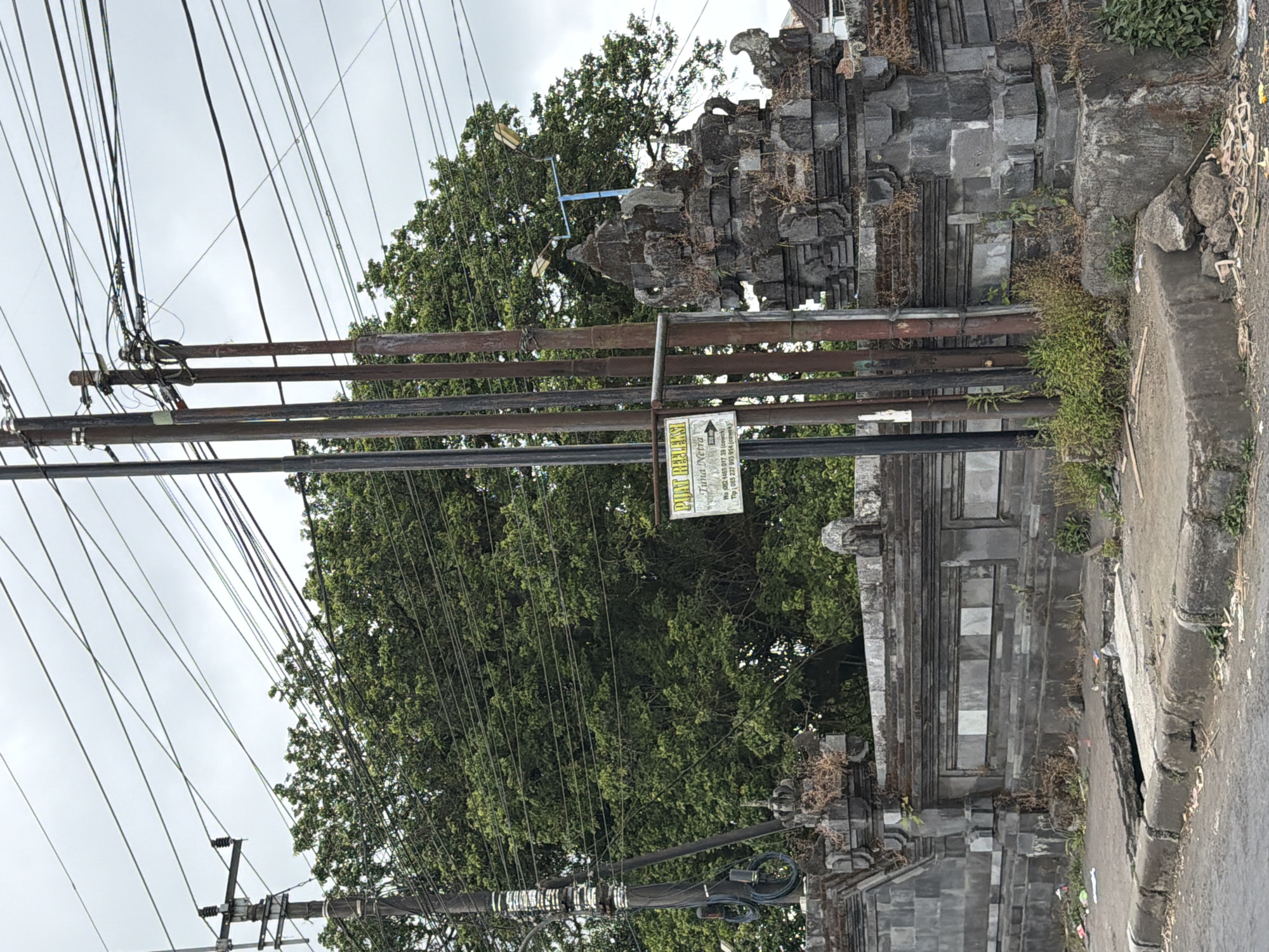 In Kintamani, Indonesia, a street scene unfolds under an overcast sky. A stone wall with intricate carvings and ornate pillars lines the foreground, leading to a street with a damaged gutter. Several utility poles, adorned with a dense network of electrical wires, stand tall beside the wall. A prominent sign, partially obscured by the poles, displays text in Indonesian and the words "PUAT REFL". To the right, a street lamp with two lights is mounted on a blue pole. Lush green trees dominate the background, their foliage creating a dense canopy. The environment appears slightly unkempt, with debris visible on the ground near the gutter and along the side of the street.