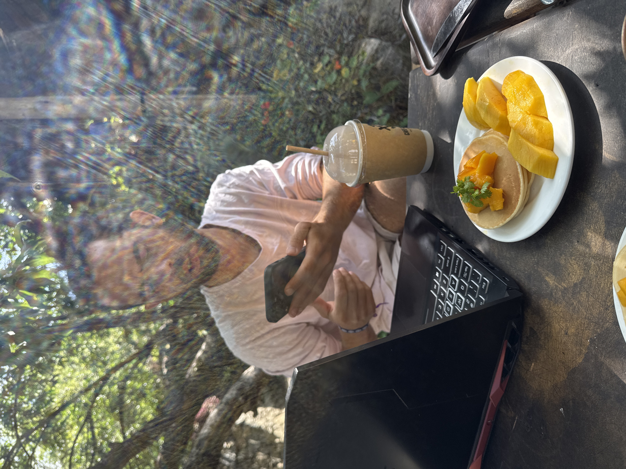 A person is seated at a wooden table outdoors, working on a black laptop. The scene is bathed in sunlight, with a rainbow-like prism effect across the top left of the image. The person is holding a black smartphone in their hands. Beside the laptop is a clear plastic cup with a straw, presumably containing a beverage. To the right of the laptop is a white plate with pancakes and sliced mango, garnished with greens. A brown leather bag sits behind the plate. In the background, through the prism effect, trees and vegetation are visible. The location is Lũng Cú Commune, Vietnam.