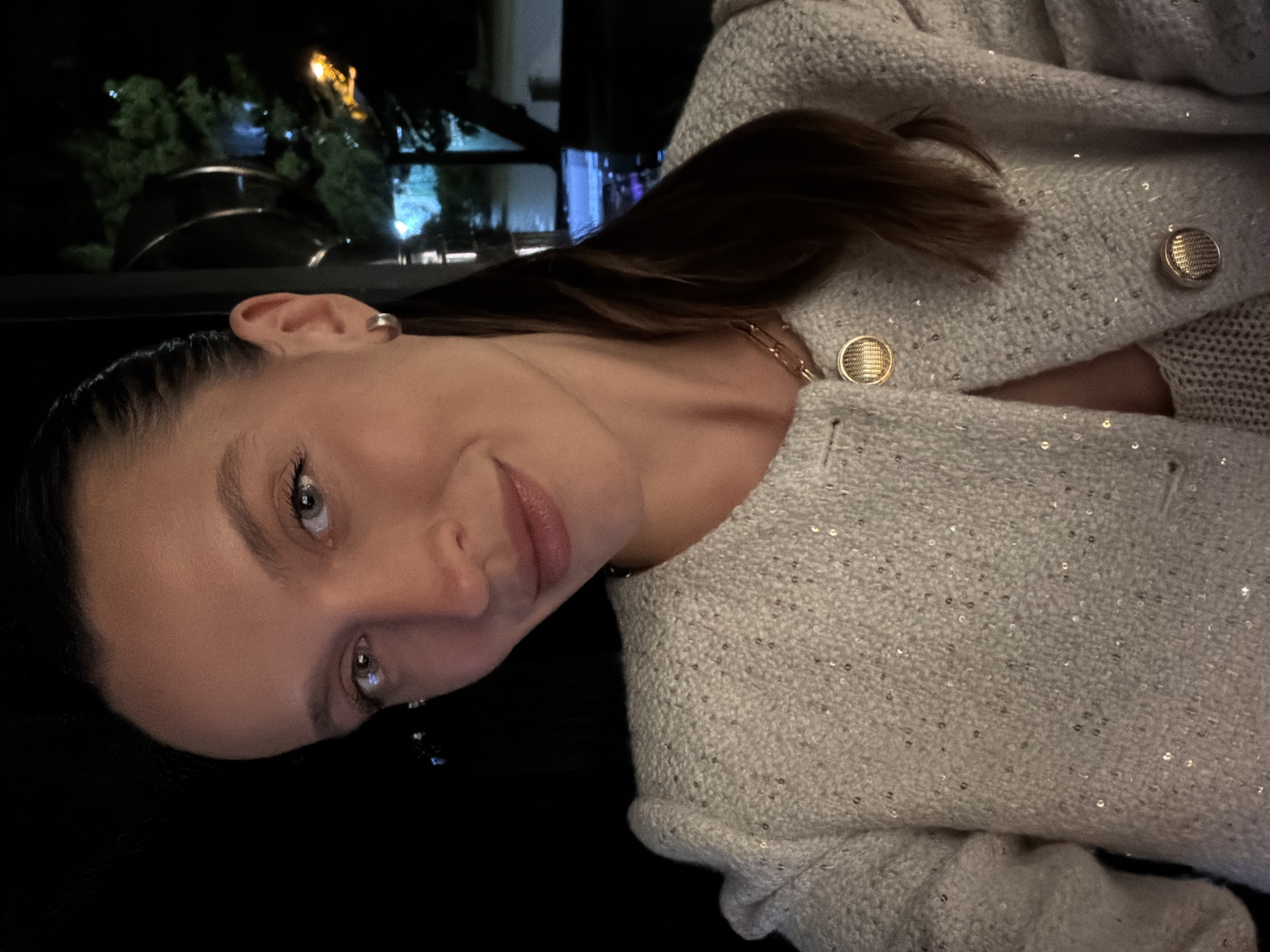 A portrait of a young woman, oriented sideways, is captured in a dimly lit setting at night. The woman looks directly at the camera with a gentle, slight smile. Her dark hair is pulled back from her face, revealing a simple silver-toned earring in her right ear. She has light-colored eyes and light pink lipstick. She is wearing a light-toned, possibly off-white or very light grey, textured jacket or cardigan. The garment is adorned with numerous small, reflective sequins and features prominent round, gold-toned buttons with a textured surface. A delicate gold chain necklace is also visible around her neck.
The background is dark and out of focus, with blurred lights and reflections, along with suggestions of dark foliage, visible through what appears to be a window or car glass. This indicates an outdoor scene at night, viewed from indoors or a vehicle. The woman's face and upper body are well-lit, contrasting with the dark surroundings. There are no specific visual cues within the image to identify the exact location, nor is any text visible. The overall mood conveyed is calm and pleasant.