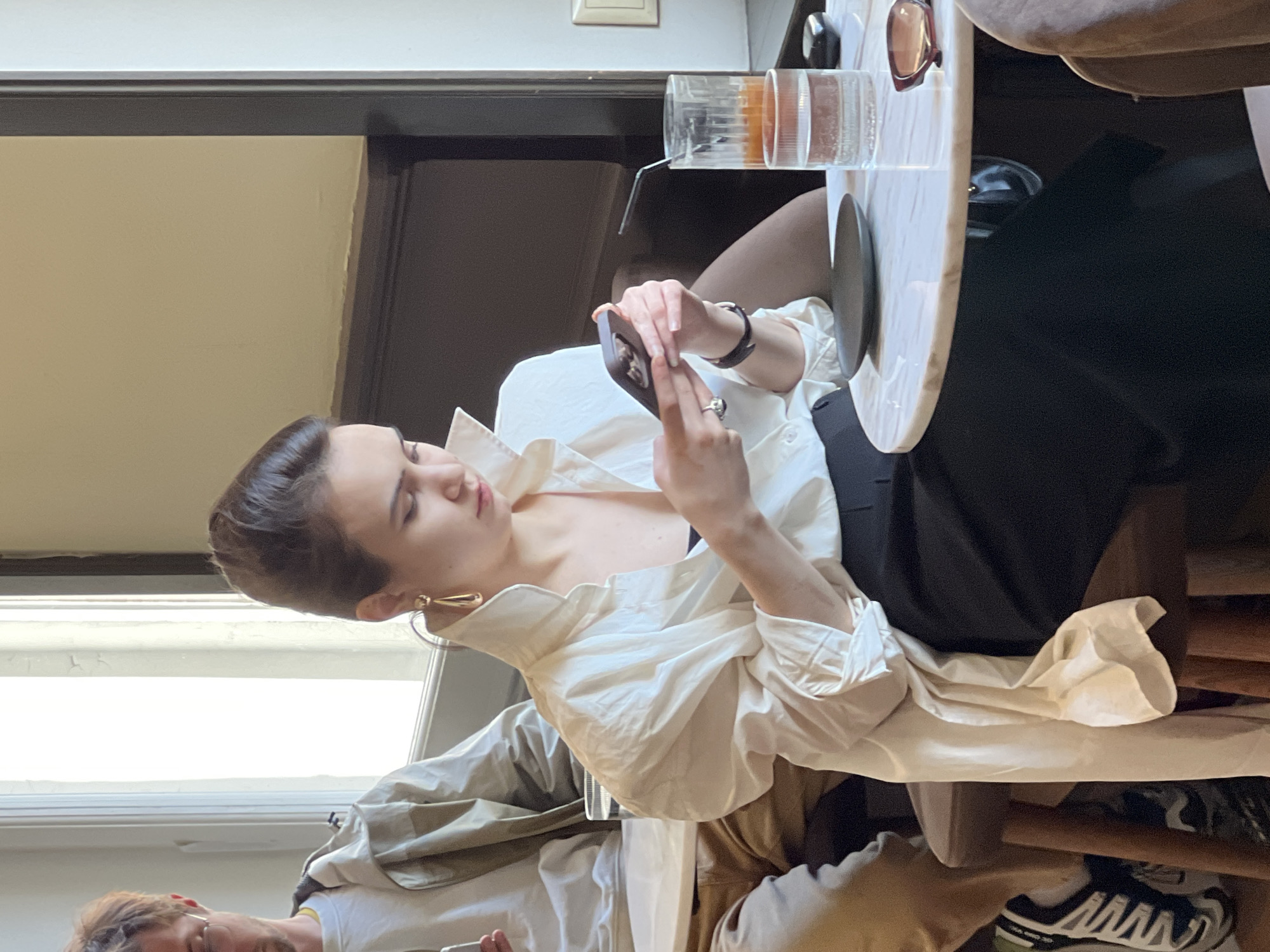 A young woman with short dark hair sits at a light-colored, round table in an indoor setting, looking at her mobile phone. She wears a voluminous white shirt and a dark skirt. A glass with a beverage sits on the table. Another person is partially visible in the lower left corner of the image. They are wearing light-colored clothing. A pair of black and white sneakers are visible under the table. Based on the interior architectural details and window style, it is potentially a restaurant or cafe in Saint Petersburg, Russia.