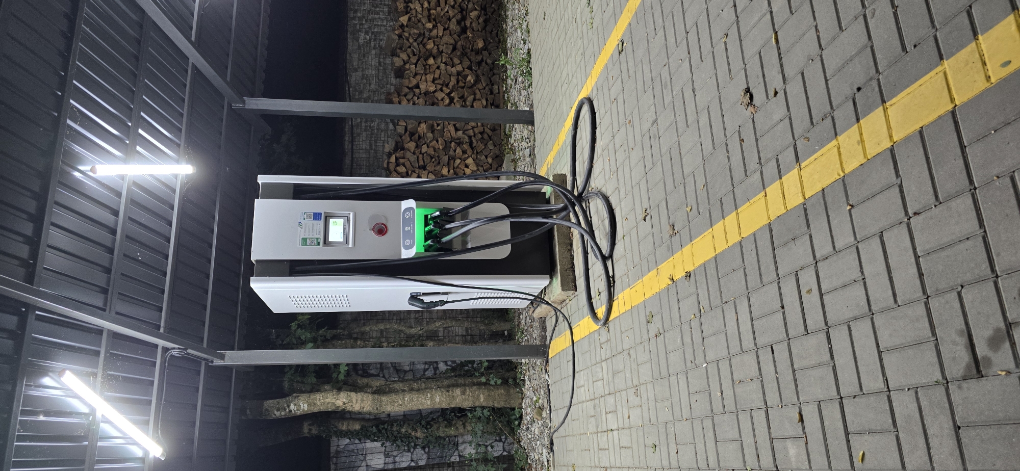 An electric vehicle charging station stands under a metal canopy with fluorescent lights. The station is white and black, with multiple charging ports that are illuminated green. Several thick black cables are connected to the charging ports and lie on the paved ground. The ground is made of gray interlocking pavers, with a yellow line marking a boundary. To the left of the charging station are trees and a stone wall covered in ivy. To the right, a neatly stacked pile of firewood is visible. The scene appears to be outdoors at night. The location is identified as Shirokaya Schel, Russia. There are no people or animals present in the image.