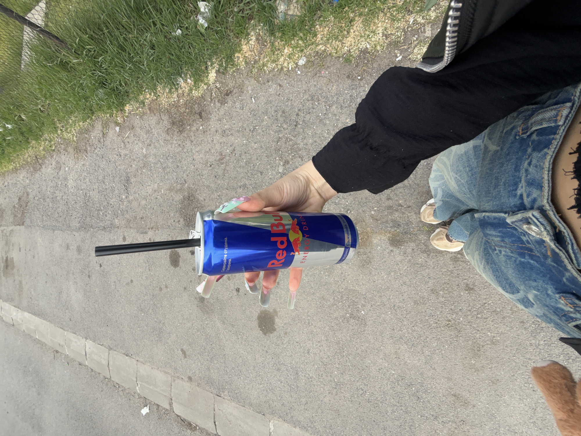 The image shows a person holding a can of Red Bull with a black straw. The person is wearing a black long-sleeved shirt and light blue jeans. The location appears to be outdoors, possibly a street or sidewalk, with grass and a fence visible in the background. The language on the can of Red Bull indicates that the image was taken in Sofia, Bulgaria.