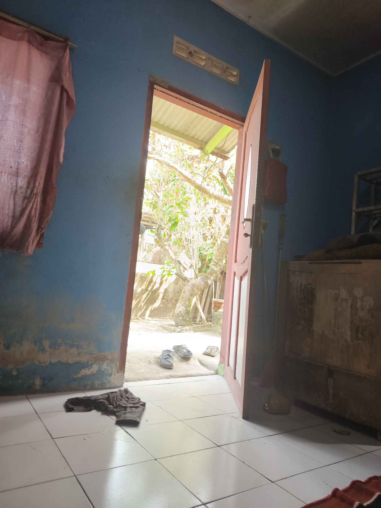 The image depicts the interior of a room in Pangkalpinang, Indonesia, with a doorway leading outside. The room has blue walls that appear weathered and stained near the floor. A pinkish, coarse-textured curtain hangs on the left side of the frame. The floor is tiled in white squares.
In the foreground, a dark-colored garment, possibly a shirt or pants, is crumpled on the floor. Just inside the open doorway, a pair of gray sandals is placed. The doorway itself is painted red and has a simple handle mechanism.
Through the doorway, an outdoor scene is visible, characterized by green foliage, tree branches, and sunlight filtering through. The ground outside appears to be a dirt or gravel pathway.
To the right of the doorway, inside the room, is a wooden cabinet or dresser, also appearing weathered. Above the cabinet, a broom or mop leans against the wall, and a red bag is hung on the door. A metal shelf unit is partially visible behind the cabinet. A small, yellow, lumpy object is on the floor near the cabinet. The ceiling is visible at the top of the frame and appears to be a suspended ceiling panel. A ventilation grate is set into the wall above the doorway.