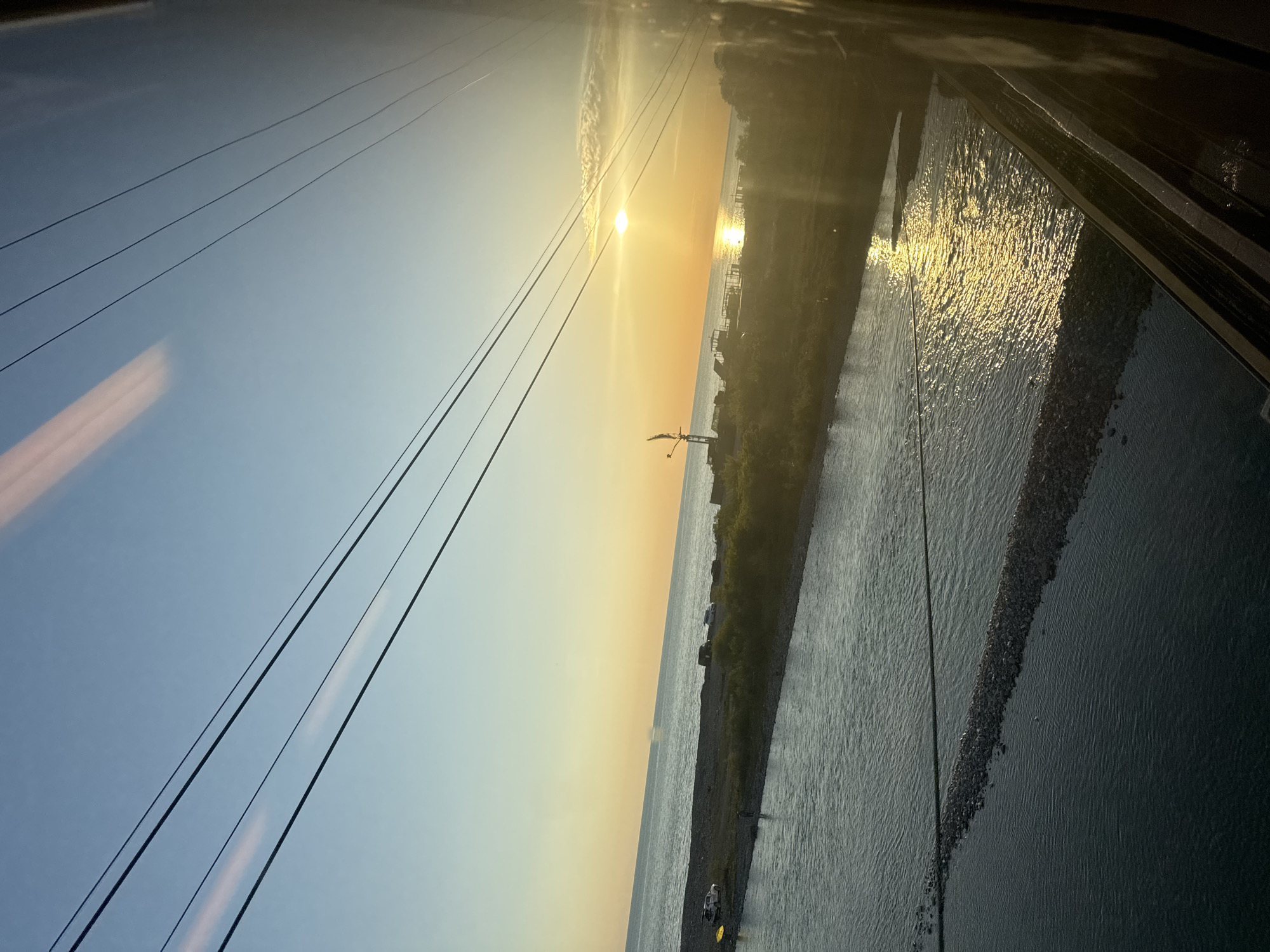 This image captures a panoramic view of the Black Sea coast in Sochi, Russia, during either sunrise or sunset. The sky, transitioning from a light blue at the top to a warm golden orange near the horizon, is dominated by a brilliant, low-hanging sun on the upper right, casting an intense, elongated reflection across the water. A thin wisp of cloud is visible above the sun.
Several parallel power lines stretch diagonally across the upper left and central portions of the sky, indicating proximity to urban or industrial infrastructure. The photo appears to have been taken from a slightly elevated position, possibly through a window, as evidenced by faint light streaks and reflections in the foreground.
The horizon line separates the sky from a vast body of water. The water directly beneath the sun exhibits strong, shimmering golden reflections. A narrow strip of land, possibly a pebble beach or embankment, runs along the coastline. On this strip, sparse vegetation and a prominent industrial structure resembling a gantry crane are silhouetted against the bright sky. Further along the coast, indistinct buildings or small structures are visible in the distance.
Closer to the foreground, the water on the right appears to be a separate, calmer body, perhaps a river or sheltered bay, bordered by a distinct, dark line of pebbles or rocks. A small, light-colored object, possibly a boat or raft with a yellow component, is seen on this calmer water section near the shore. No people or visible text are discernible in the image. The scene conveys a sense of serene natural beauty combined with subtle hints of human development.