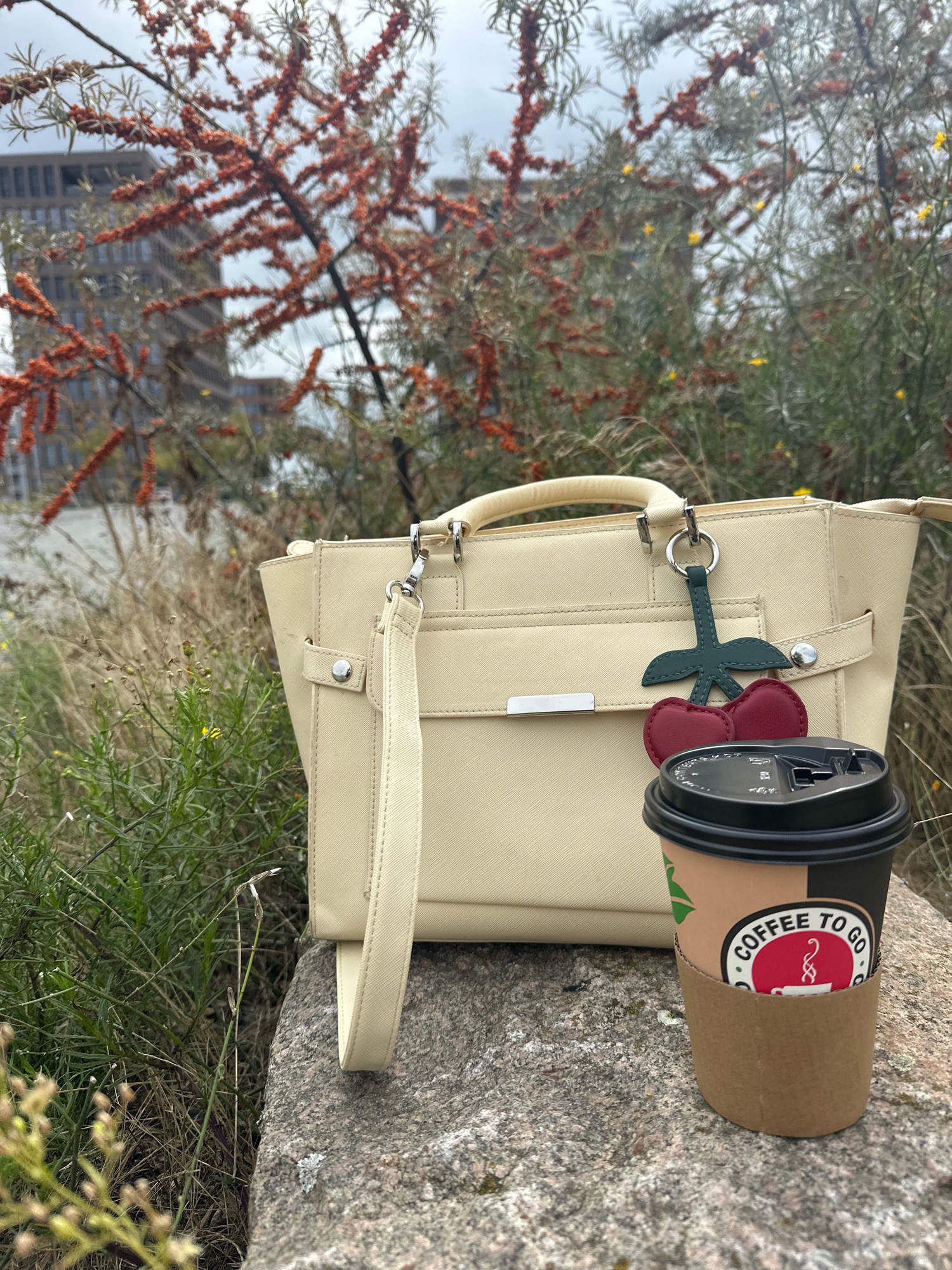 A cream-colored tote bag with a cherry charm and a disposable coffee cup sit on a stone wall. The bag has a top handle and a detachable shoulder strap, with a silver buckle detail on the front. The coffee cup is brown with a black lid and has a sticker that says "COFFEE TO GO". In the background, there are trees with red berries and buildings. The scene appears to be outdoors in an urban setting, possibly in Bremen, Germany, with a cloudy sky overhead.