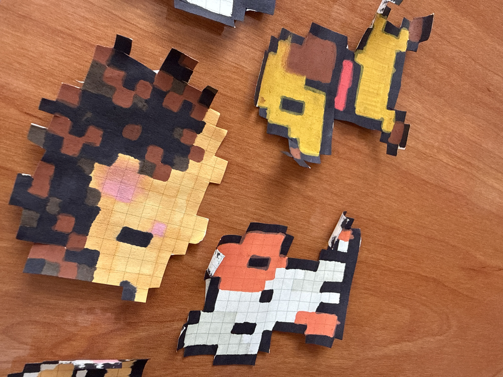The image shows several paper cutouts with pixelated designs on a wooden surface. The cutouts appear to be animals rendered in an 8-bit style. One resembles a dog with yellow and brown coloring. Another looks like a person with black hair. The location in the city of Unknown, Unknown is irrelevant to the visual content of the image.