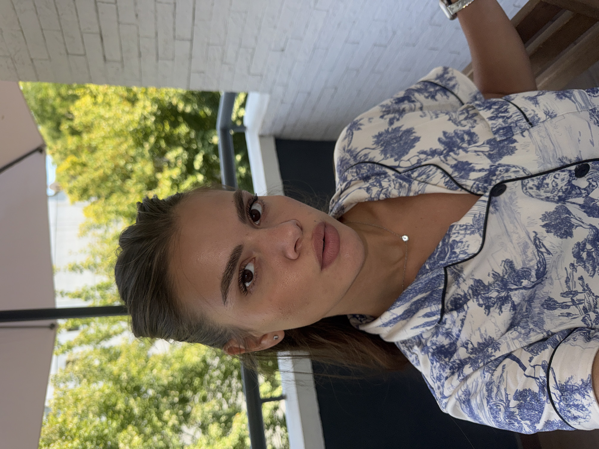 A young woman with brown hair pulled back and wearing a blue and white patterned pajama top is captured in a close-up, outdoor shot. She is looking directly at the camera with a neutral expression. Her makeup is subtly applied, enhancing her features. She wears a small, delicate silver necklace with a single clear pendant. Her left earlobe has a small stud earring. The background features lush green trees, a white brick wall, and a dark railing, suggesting she is on a balcony or terrace. The lighting indicates it is daytime, possibly late morning or early afternoon, with soft sunlight filtering through the leaves. The environment suggests a peaceful, residential setting, possibly in a city area with green spaces.
