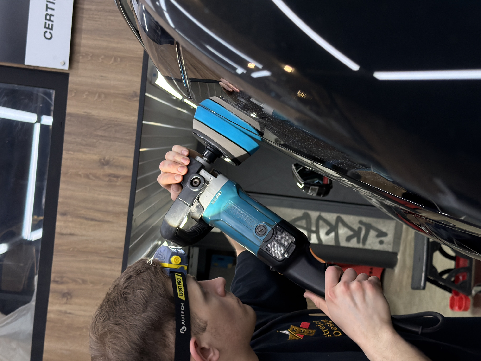A person is polishing a black car in a garage. The individual wears a black shirt with a logo, a headlamp marked "AUTECH LIGHT PRO," and holds a blue and gray polishing tool against the vehicle's surface. The garage setting includes wood paneling on the walls, lighting fixtures, and a glimpse of graffiti or signage in the background. There is no indication in the image that this location is in Krasnoyarsk, Russia.
