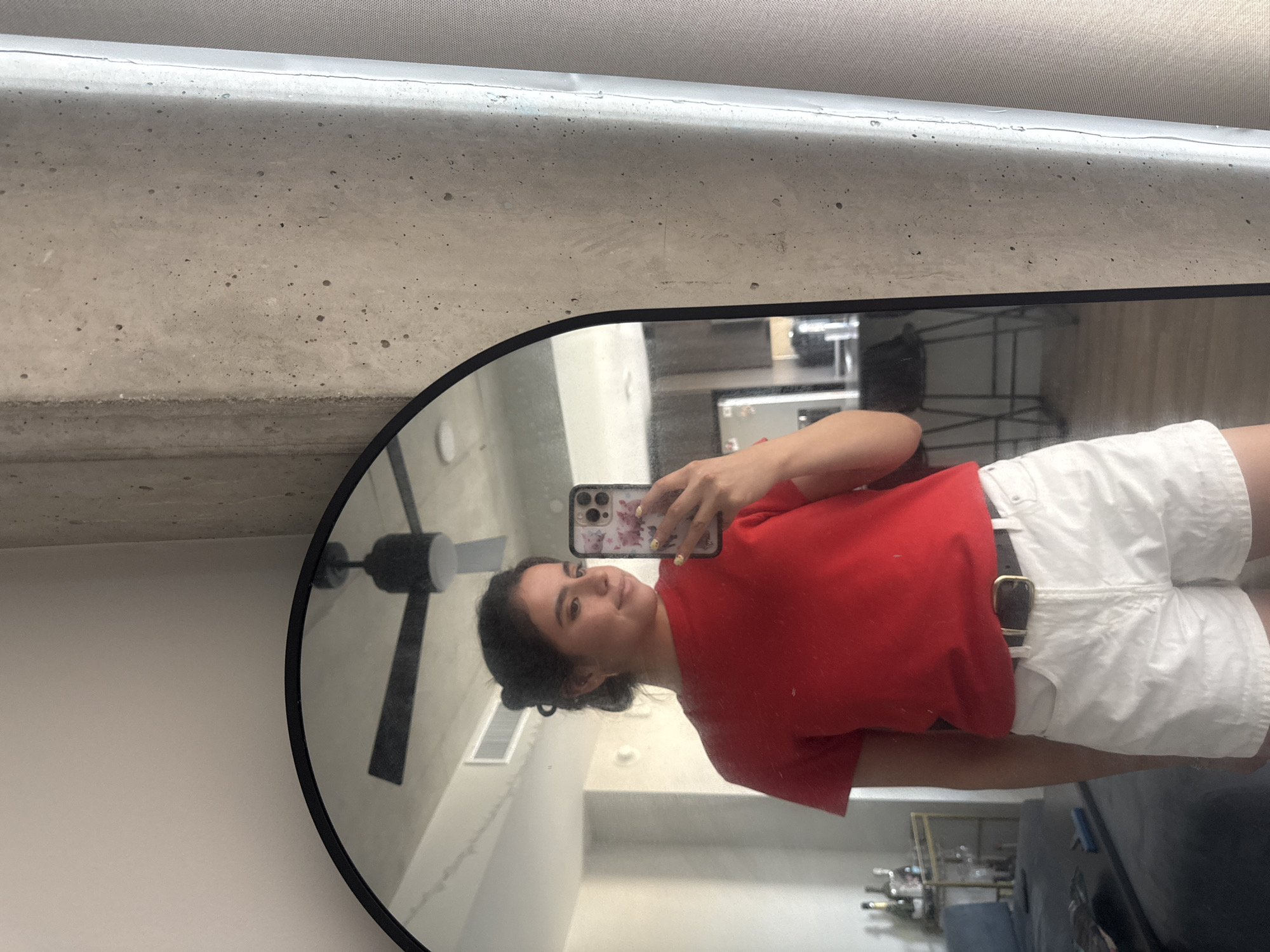 A woman is taking a mirror selfie indoors, likely in her apartment. She is wearing a red shirt and white shorts with a black belt. The room appears to be modern, with a visible ceiling fan and kitchen area with stainless steel appliances. The location is indicated as Austin, United States of America, but there are no specific landmarks to confirm this.