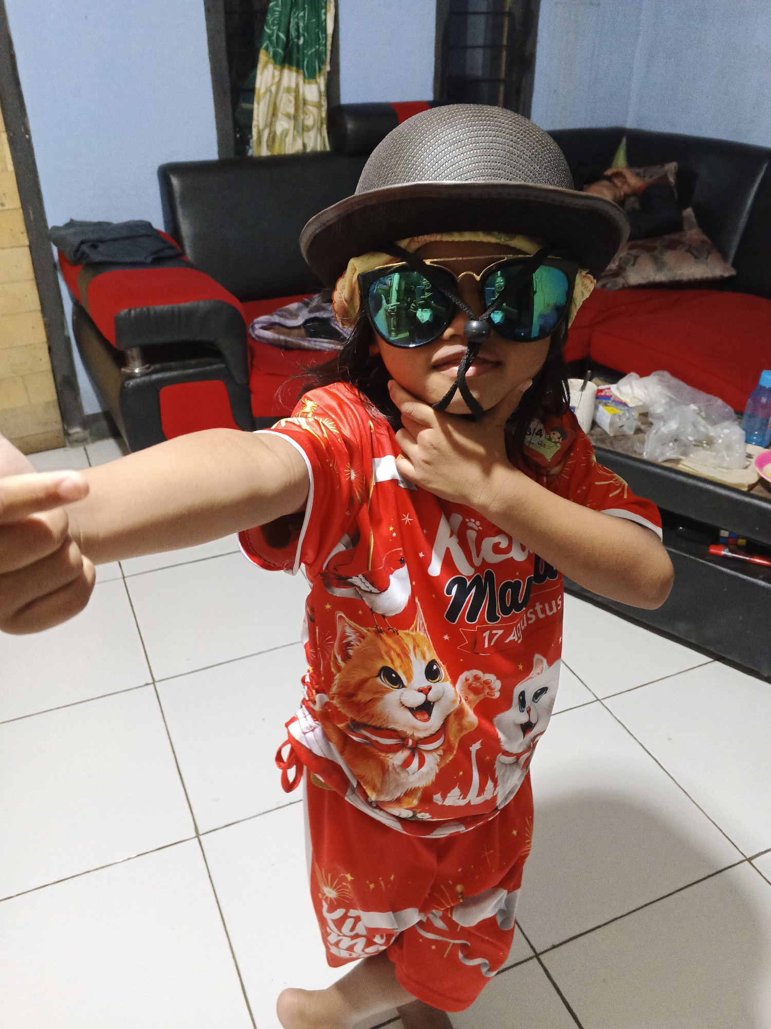 A child, appearing to be around four to six years old, stands in what seems to be a domestic indoor setting. The child is wearing a red t-shirt and shorts adorned with cartoon cat designs and celebratory text. They are also sporting a grey hat, oversized green sunglasses with reflections of onlookers, and a black accessory across their nose. The child's left arm is extended forward, with their index finger pointing. The background includes a black leather sofa with red accents, a tiled floor, and some indistinct objects on a shelf. The lighting suggests it is daytime.