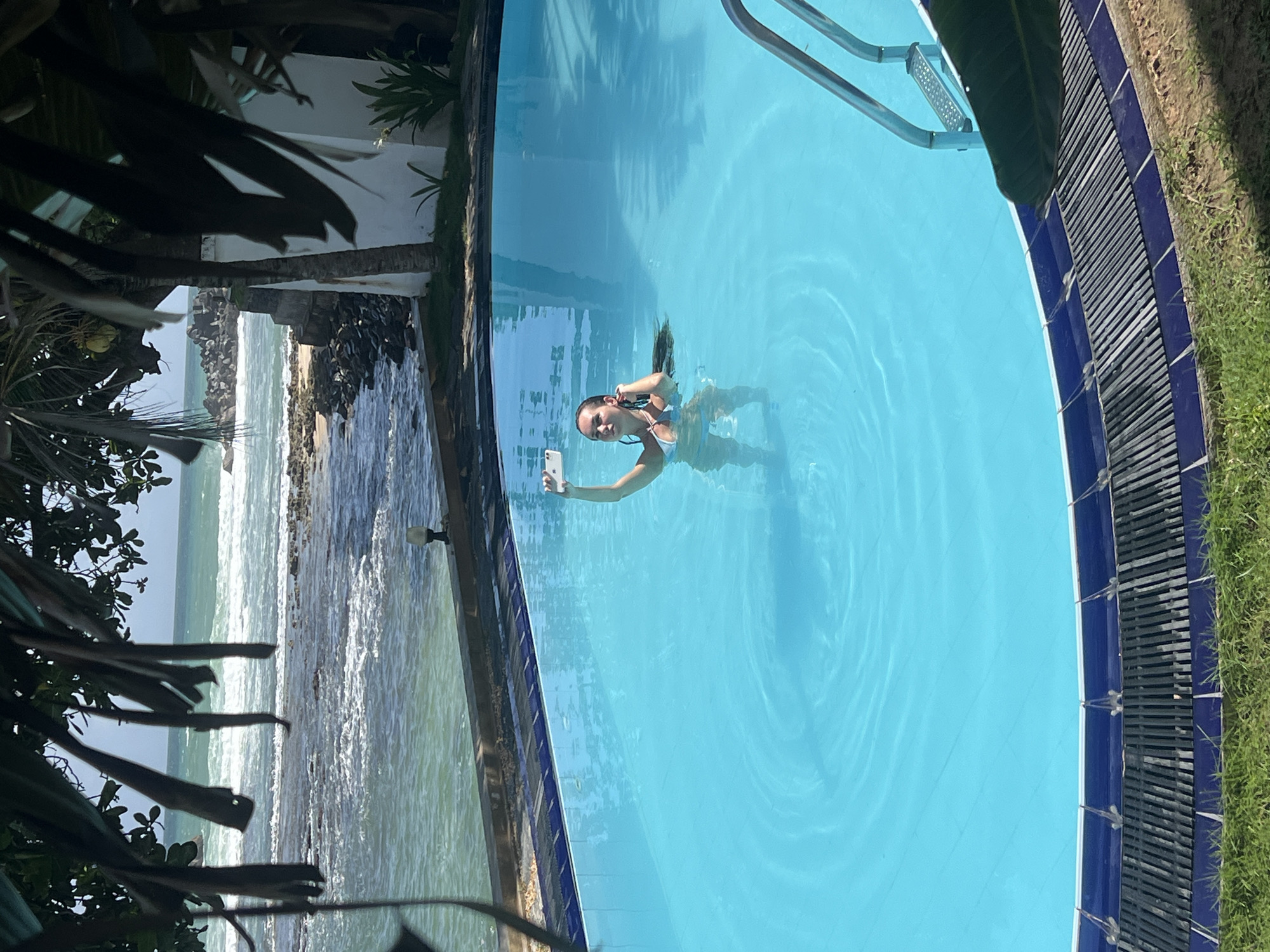 A young woman in a light-colored bikini stands in the middle of a bright blue swimming pool, taking a selfie with her smartphone. She has long, dark hair, some of which is submerged in the water. The pool is bordered by blue tiles and a dark grate, with green grass visible beyond the pool. In the background, the turquoise ocean can be seen with waves breaking on a rocky shore. Palm leaves hang from above, partially obscuring the view of the sky and a white building with a palm tree in front of it. The location is Goviyapana, Sri Lanka.