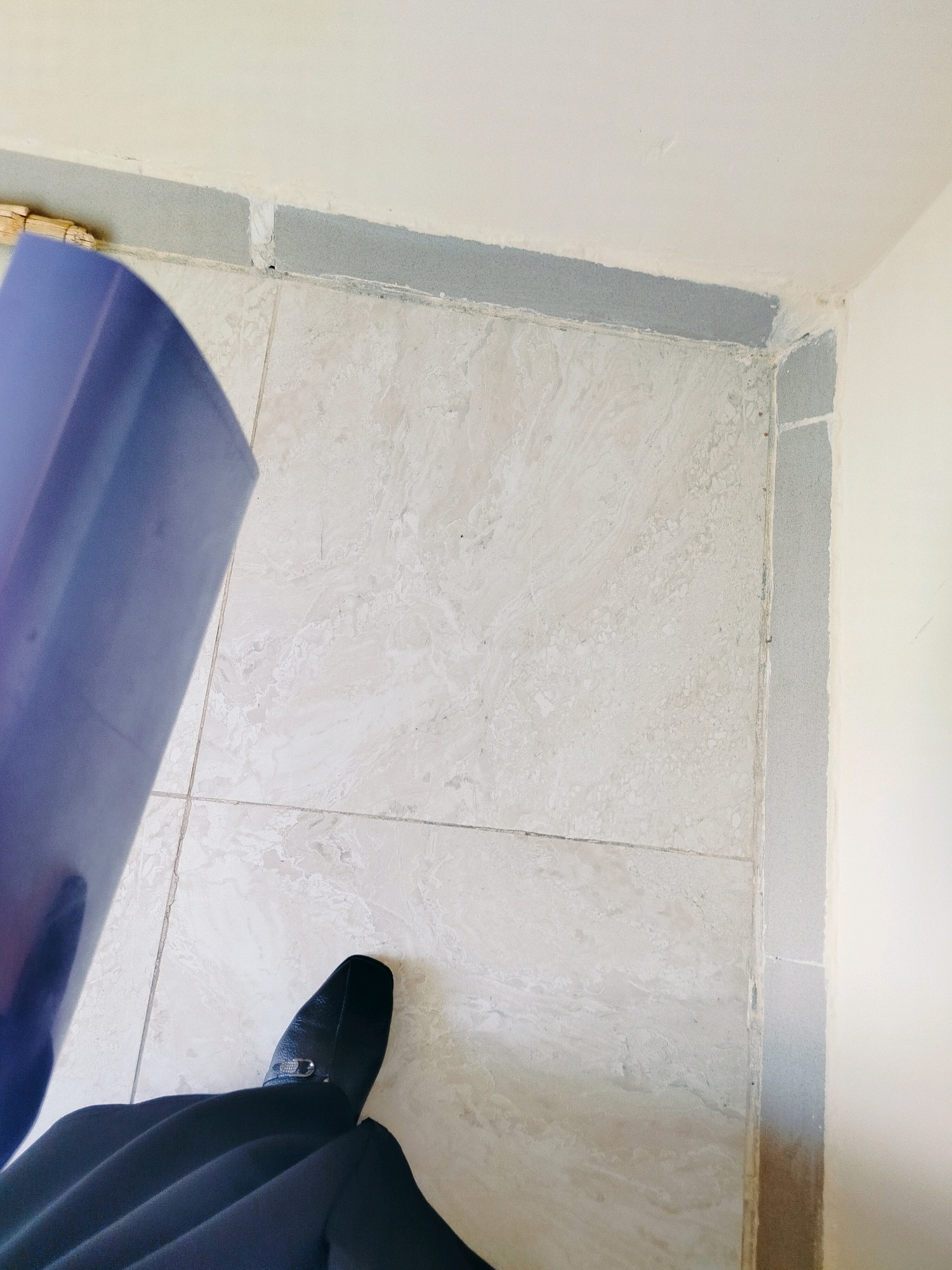 The image shows a close-up view of a tiled floor and the corner of a room with unfinished walls in Көлкент, Kazakhstan. A person's black shoe and the dark fabric of their pants are visible in the foreground. A large, purple object, possibly a rolled-up tarp or fabric, is held by an unseen person, casting a shadow on the floor. The tiled floor is a light beige with subtle grey veining, and it appears to be laid in large square tiles. The walls are a light cream color, and there is a grey band of construction material along the base of the walls where they meet the floor. The scene suggests a construction or renovation site.