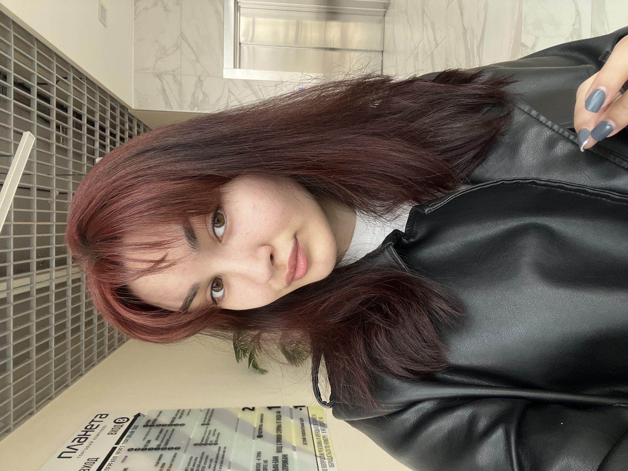 Here's a detailed description of the image:
A young woman with reddish-brown hair and bangs is posing for a close-up selfie. She has light skin and brown eyes. She is wearing a black leather jacket over a white top. Her nails are painted a matte gray color.
In the background, there is a wall with a grid-like pattern and a section of marble-like tiles. A sign is partially visible at the bottom of the frame with Russian text, the text "планета" is visible which translates to "planet", indicating a shopping mall or complex. The sign also lists various commercial establishments. Based on the sign and the text, the location is likely Neftekamsk, Russia.