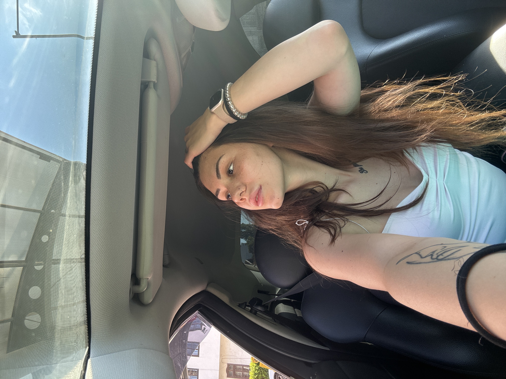 A young woman with long brown hair sits in the passenger seat of a car, looking out the window. She is wearing a white tank top and has several tattoos visible on her arm and chest. She is also wearing a smartwatch and bracelets on her left wrist. The interior of the car is visible, including the black leather seats and the car's sunroof. Outside the car, a building with windows is visible, along with a pole and trees. The sky is blue with some clouds. The image appears to have been taken in Sofia, Bulgaria, based on the architectural style of the building.