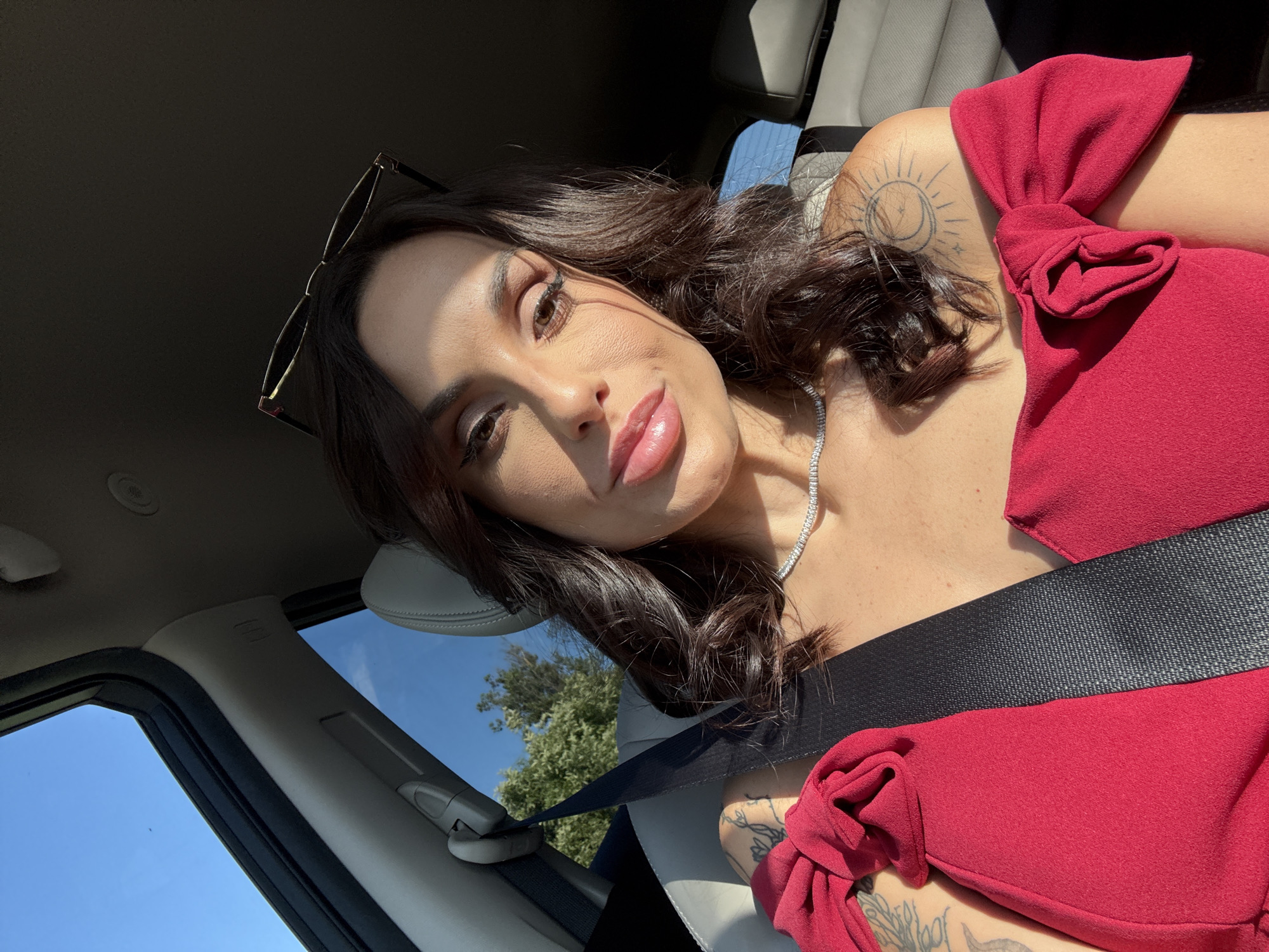 A woman with medium-length brown hair, wearing a red top and a silver necklace, is seated in a car. She is wearing a black seatbelt. Sunglasses are perched on her head. She has tattoos on her shoulder and arm. The car interior is light-colored. Trees and blue sky are visible through the window. The location is unknown, the information about Kaleytsa, Bulgaria is incorrect.