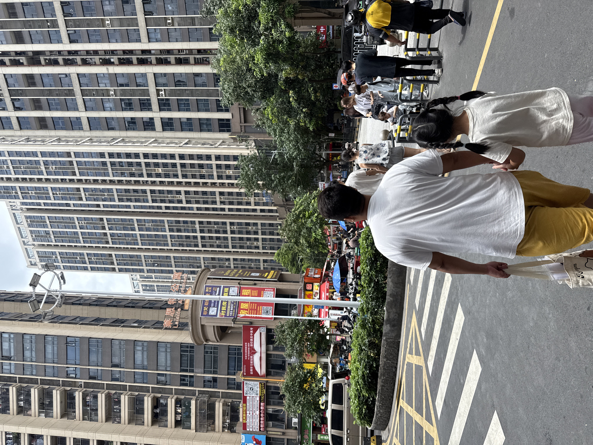 In Haizhu District, China, a father and daughter walk hand-in-hand across a pedestrian crossing. The father, wearing a white t-shirt and yellow shorts, carries a tote bag. The daughter, with braided pigtails, is dressed in a white t-shirt and pink pants. They are surrounded by tall apartment buildings, a street light, and lush green trees. The area is busy with other pedestrians, vehicles, and market stalls. The sky is overcast.