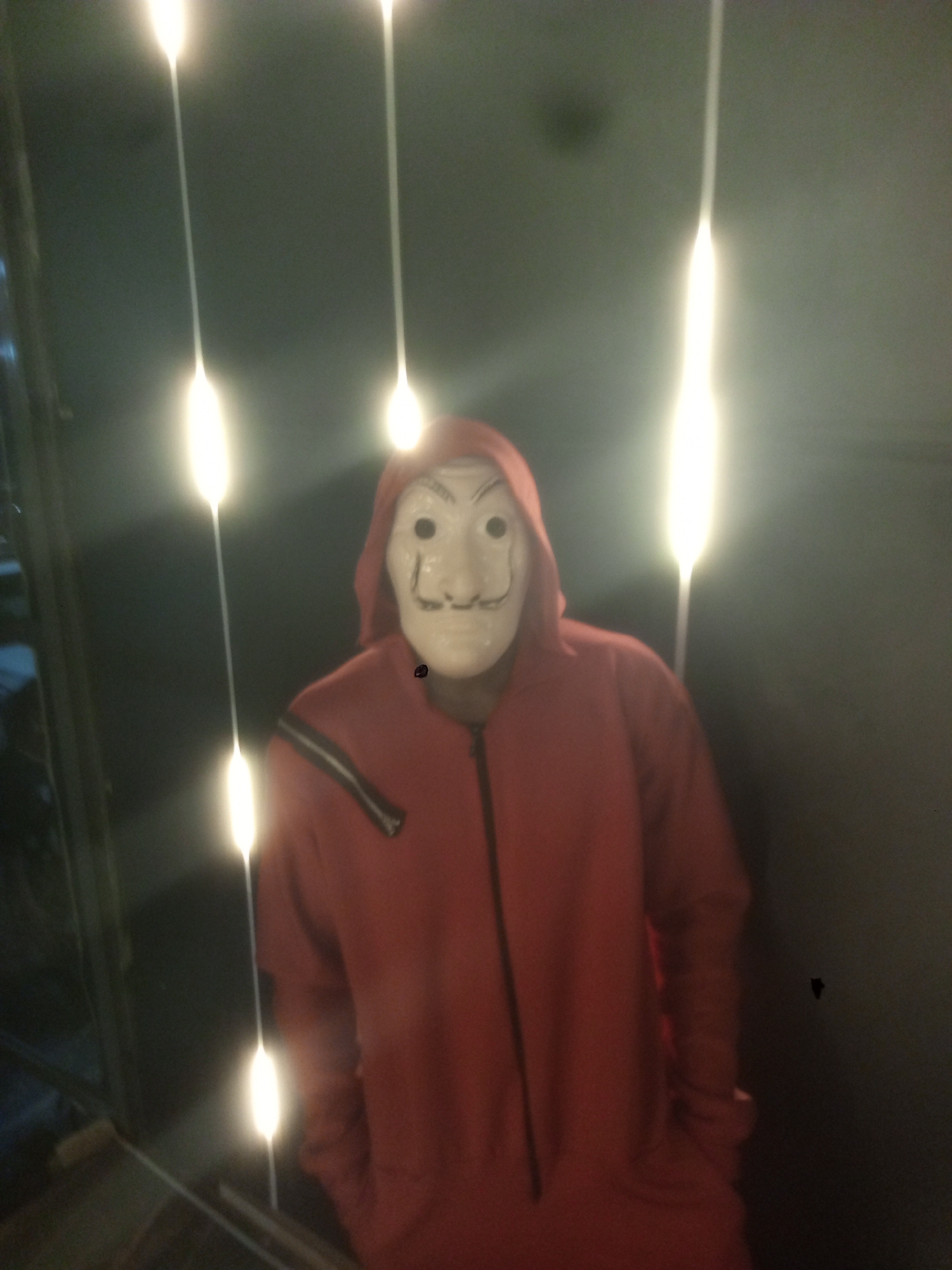 A person wearing a red jumpsuit and a Salvador Dali mask stands in front of a wall with vertical strings of lights. The location is Gwarinpa, Nigeria. The person's hands are in their pockets. A zipper is visible on the jumpsuit. There is a blemish on the wall to the right of the person.