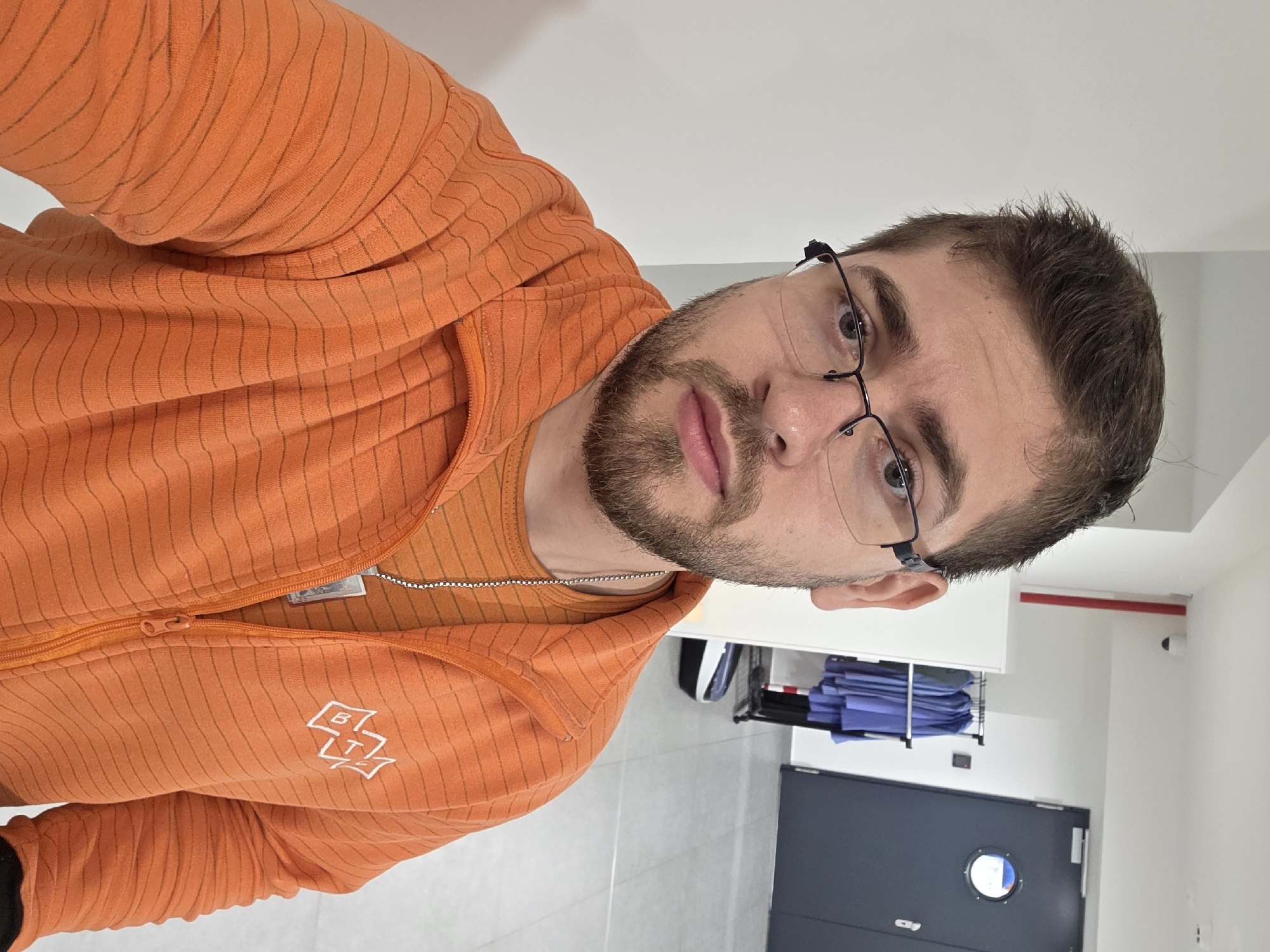 A man with brown hair, a beard, and glasses is posing for a selfie indoors. He is wearing an orange hoodie and an orange t-shirt. In the background is a room with white walls, a gray door with a circular window, and medical scrubs stacked on a rack. There is no indication of Plovdiv, Bulgaria in the image.