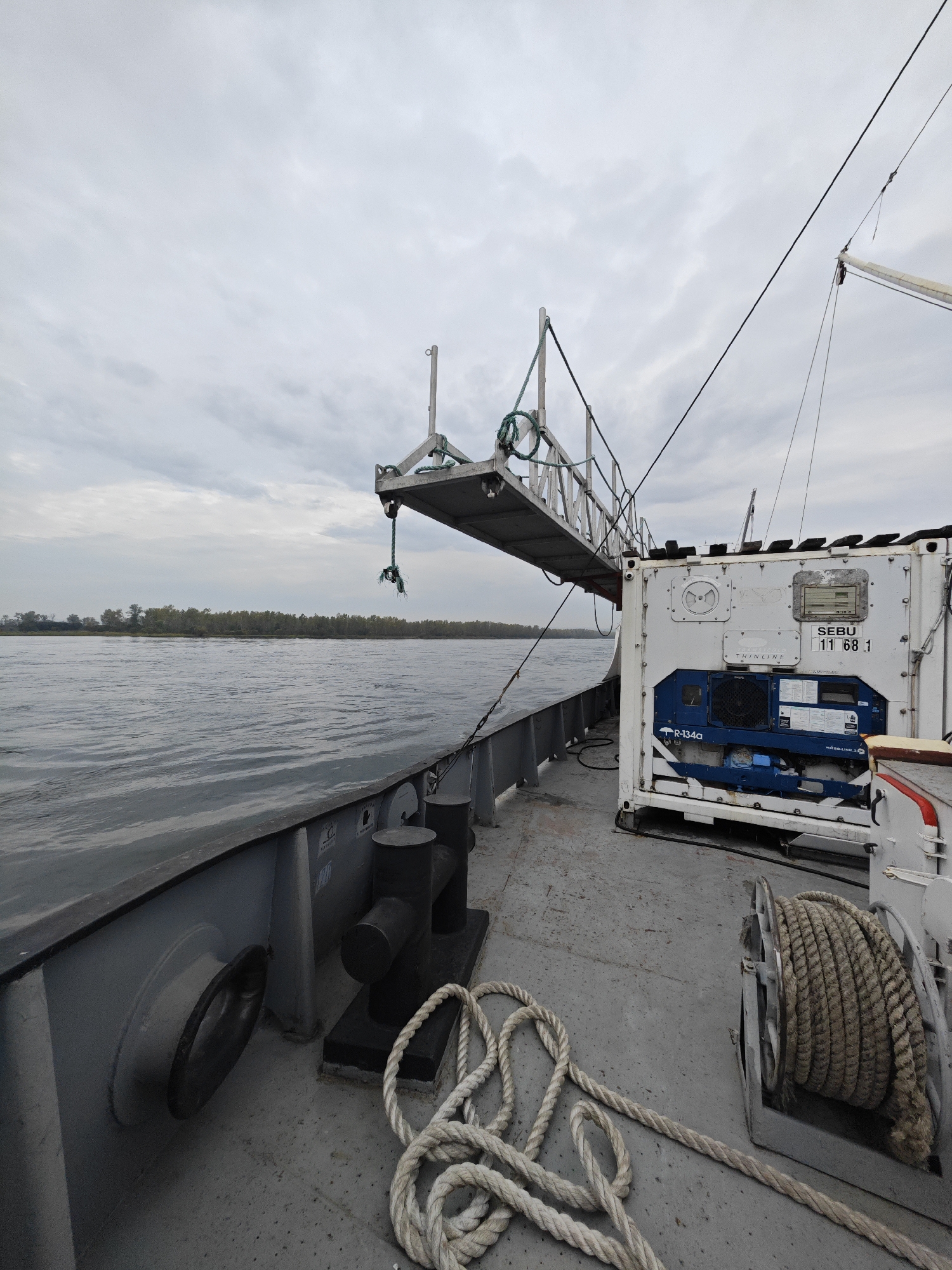 The image shows the deck of a boat on a river, with a cloudy sky overhead and trees on the distant shore. In the foreground, a large white refrigerated container is visible, with the text "SEBU 11 68 1" printed on it. A metal gangway extends from the boat towards the shore, with a rope attached to it. A coiled rope lies on the deck near the container. Mooring bollards are also present on the deck. The scene is set in Ермолаево, Russia.
