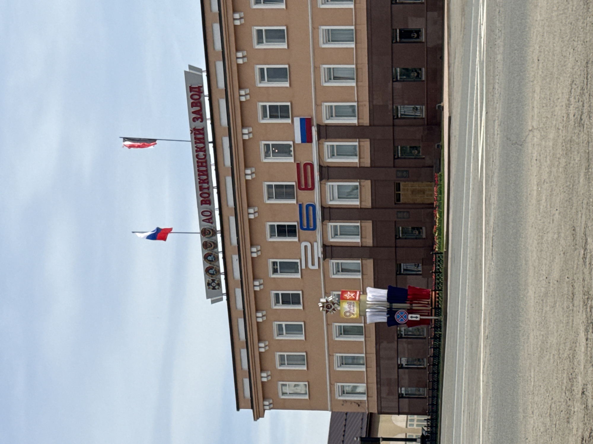 The image depicts the administrative building of the Votkinsk Plant in Votkinsk, Russia. The building is multi-story and of mixed construction, primarily light brown on the left and dark brown on the right, with numerous windows. The name of the company, "Votkinsk Plant" is displayed vertically along a sign on the left side of the building. Two Russian flags are displayed on poles. Below the company name sign, a circular sign depicting portraits and medals is present. The number "260" is displayed in large red and blue numbers on the building facade. Near the base of the building are several banners and signs arranged in a small display. The image also contains a view of a gray asphalt street that is partially visible. The sky is pale blue.