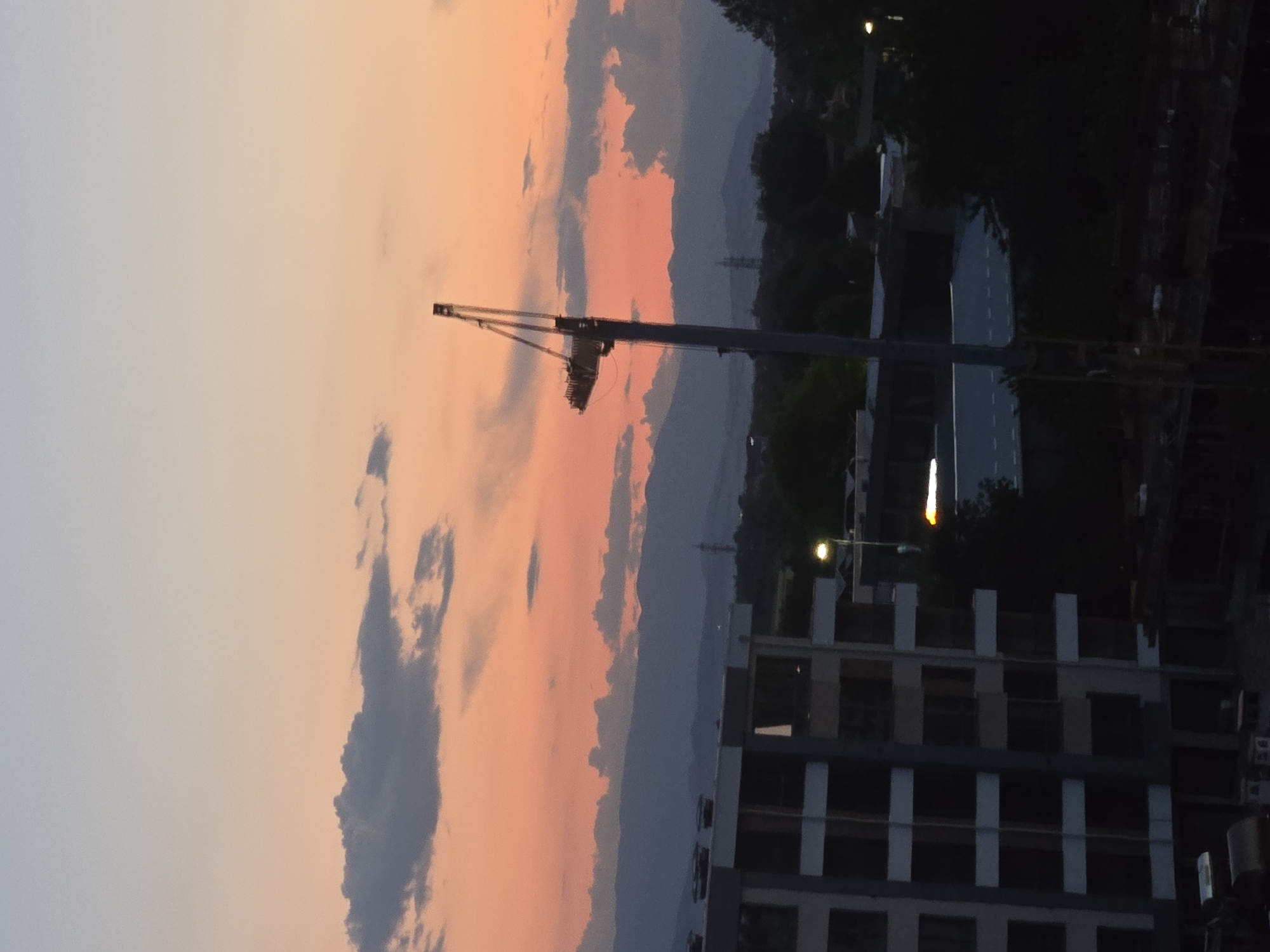Here's a concise description of the image:
The image depicts an urban scene at dusk, dominated by a construction crane silhouetted against a sunset sky. The sky is colored with shades of orange and grey clouds. A multi-story building, seemingly under construction or recently completed, is visible in the lower right of the frame. A road and streetlights are seen to the right of the building, behind trees. The location is the city of Unknown, Unknown.