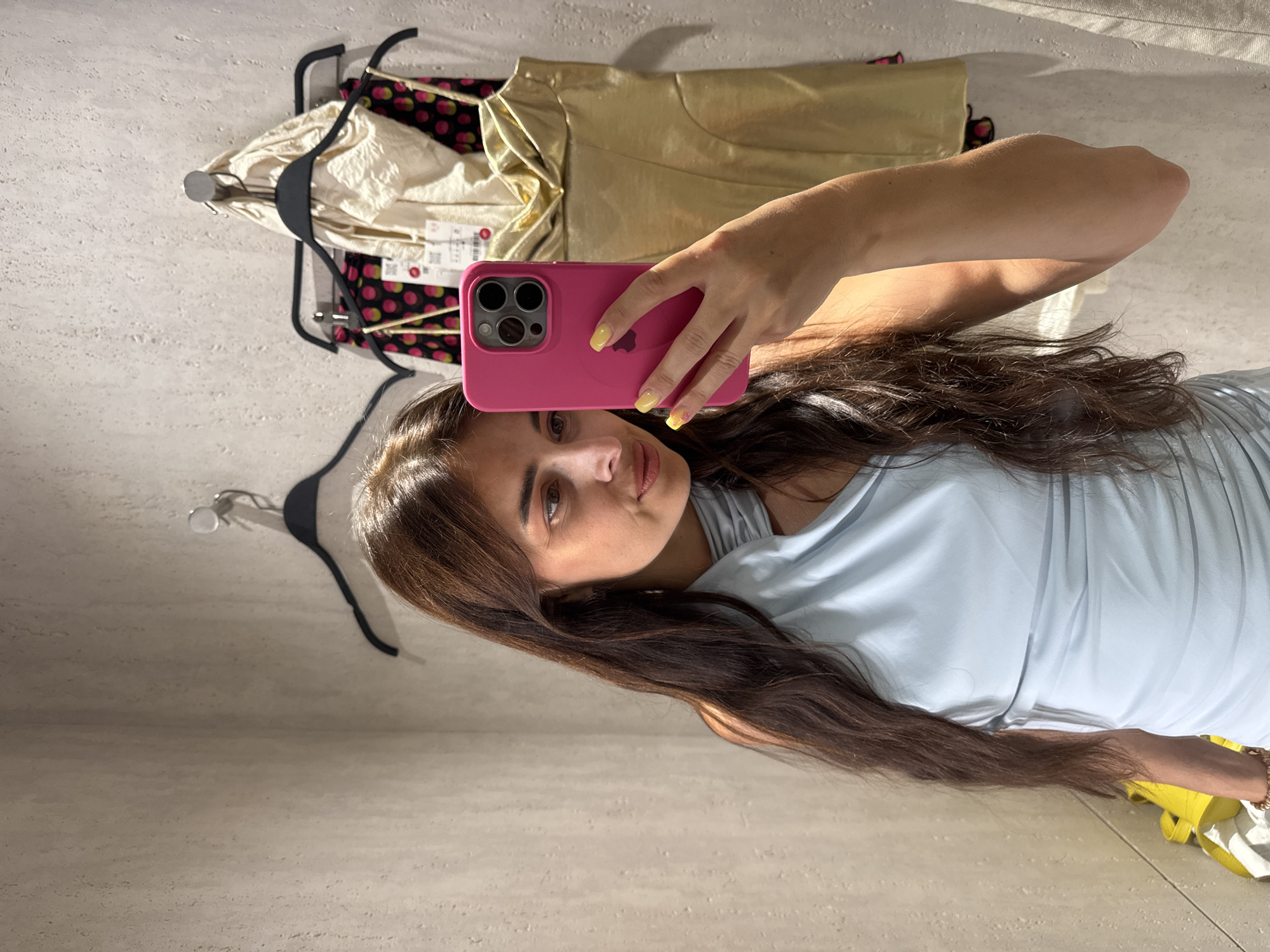 A young woman with long, dark brown hair takes a mirror selfie in what appears to be a fitting room. She is holding a pink smartphone up to take the photo. She is wearing a light blue, off-the-shoulder top. In the background, several garments are hanging on a rack, including a cream-colored top, a black top with pink polka dots, and a shimmering gold garment. The fitting room walls have a textured, light-colored finish. The lighting suggests it is daytime, possibly with natural light casting shadows. The woman's expression is neutral as she focuses on taking the selfie. There are no visible signs or text in the image.