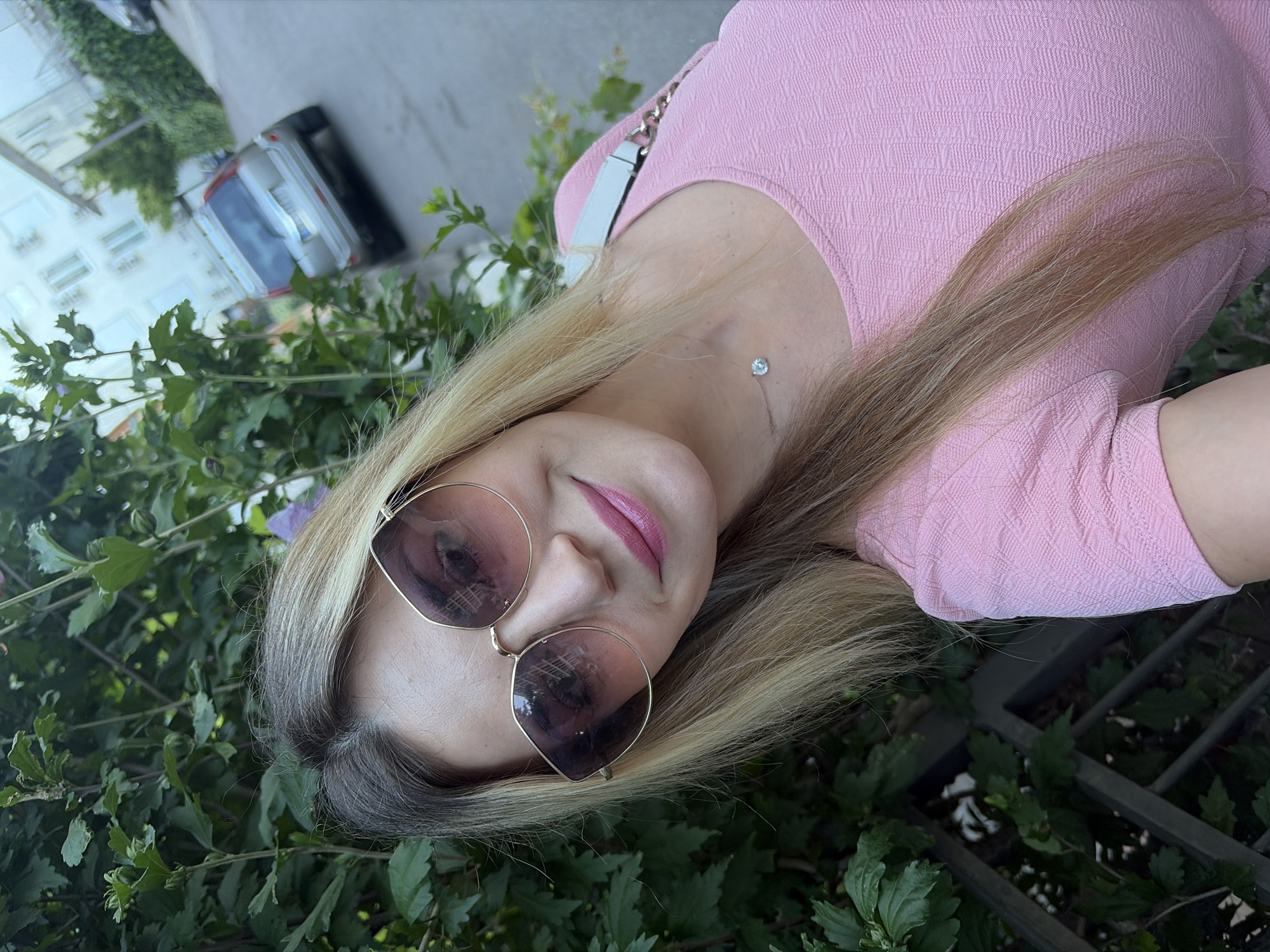 In Budva, Montenegro, a young blonde woman with sunglasses and pink lipstick smiles at the camera. She is wearing a pink textured dress and a delicate necklace with a single stone. The background features lush green foliage, a paved road, and a silver car parked on the side. Buildings are visible in the distance. The image is a selfie taken outdoors on a sunny day.