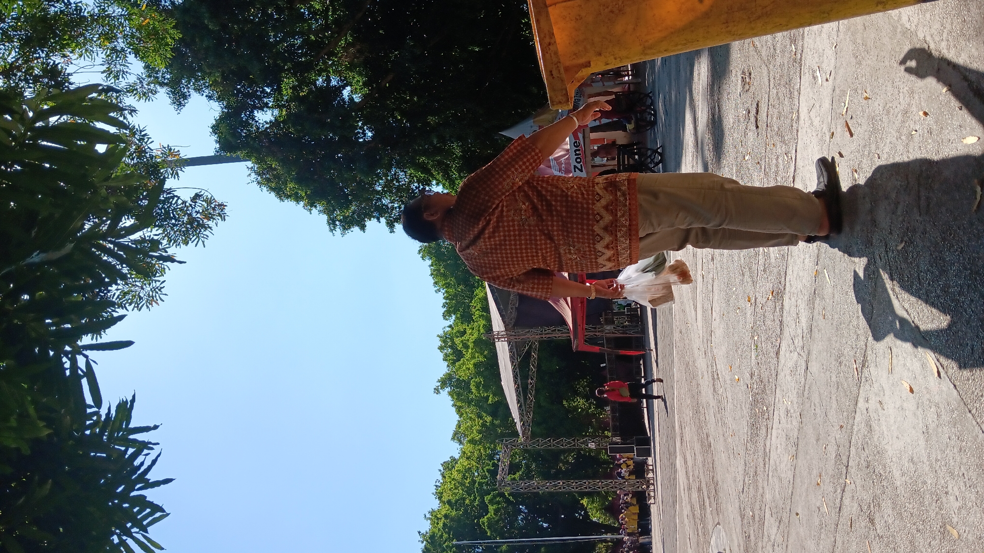 In Nganjuk, Indonesia, on a clear, sunny day, a man in a patterned collared shirt and tan pants walks away from the camera, holding a plastic bag with food. He is near a large yellow trash receptacle and a stage set up outdoors, with trees and foliage framing the scene. In the background, a crowd gathers near the stage, suggesting an event or performance is taking place. The ground is paved, and fallen leaves are scattered across its surface.