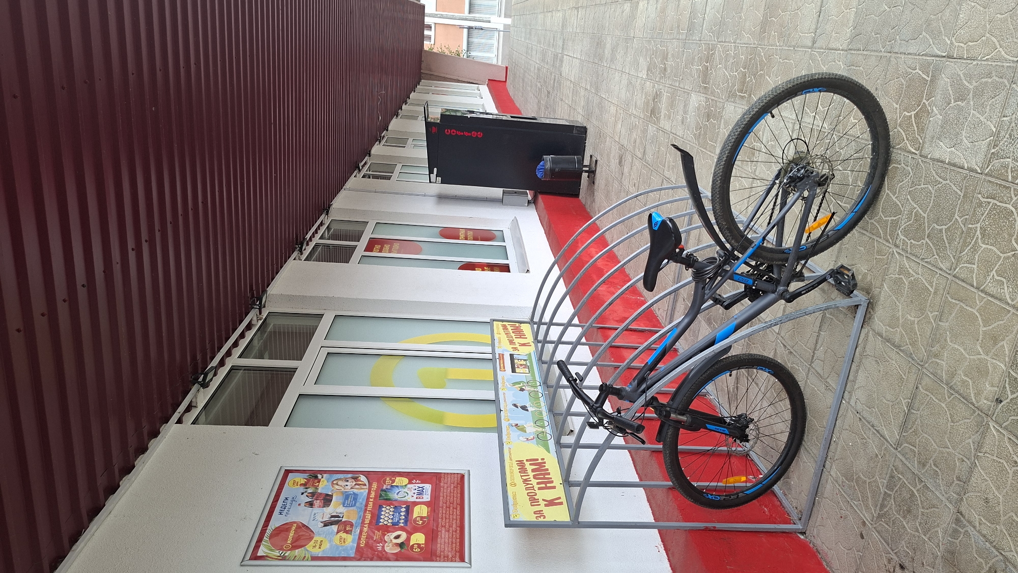 The image depicts a bicycle parked at a bike rack outside a building. The bike is dark gray with blue accents on the rims and frame. It is a modern mountain bike with front and rear suspension, disc brakes, and knobby tires. The bike rack is made of metal, painted gray, and has a curved design to hold multiple bicycles. Above the bike rack, there is a sign with text in Russian, which translates to "FOR PRODUCTS TO US!". To the left of the bike rack, a poster is displayed on the building's wall, featuring various products and promotional offers. The building itself has a white facade with large windows and a dark red corrugated roof. The ground is paved with square tiles. In the background, a black vending machine or cabinet is visible, and further down the sidewalk, there are more windows and sections of the building. The scene appears to be an outdoor area in a city or town. It is not possible to determine the specific location within Myadzyel, Belarus from this image alone.