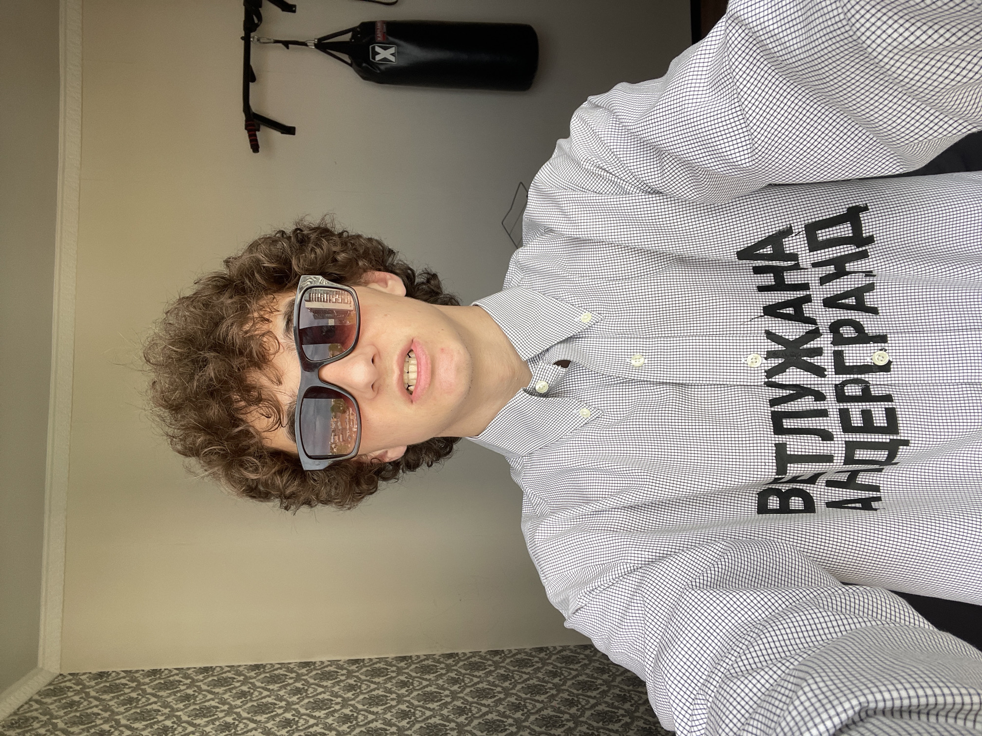 A man with curly brown hair and sunglasses is wearing a white and navy checkered button-down shirt with black Cyrillic text that appears to say "ВТЛУЖАНА АНДЕРГРАУНД" ("Vtluzhana Underground"). In the background, there is a wall and a black punching bag with a white "X" logo, hanging from a black frame. The sunglasses reflect the image of a building and trees, possibly indicating an outdoor location. The floor has a patterned carpet. The photo is taken at an angle, suggesting the camera was held at a tilted position. The location in Krasnoyarsk, Russia is only supported by the text on the shirt.