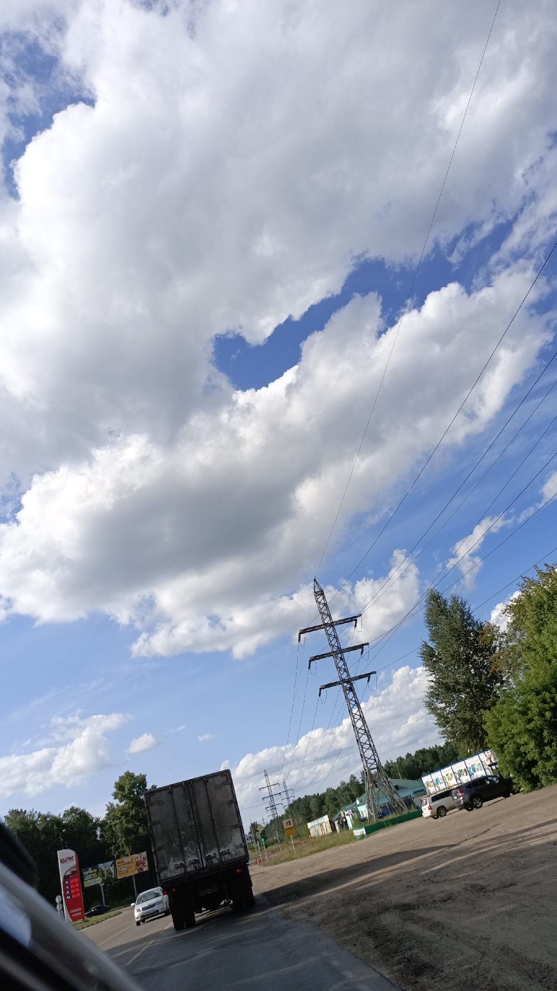 The image shows a view of a road with a truck and cars. There are power lines and towers in the background, as well as trees and buildings. The sky is blue with white clouds. The location is in Angarsk, Russia. The scene appears to be a sunny day with good visibility. There are no people visible in the image. The truck is a large, dark-colored vehicle, and it is in motion. There are several cars on the road as well. The power lines are strung between tall towers, and they are a prominent feature of the landscape. The trees are green and lush, and they appear to be in a wooded area. The buildings are a mix of structures, some appearing to be commercial. The road is paved and appears to be in good condition.