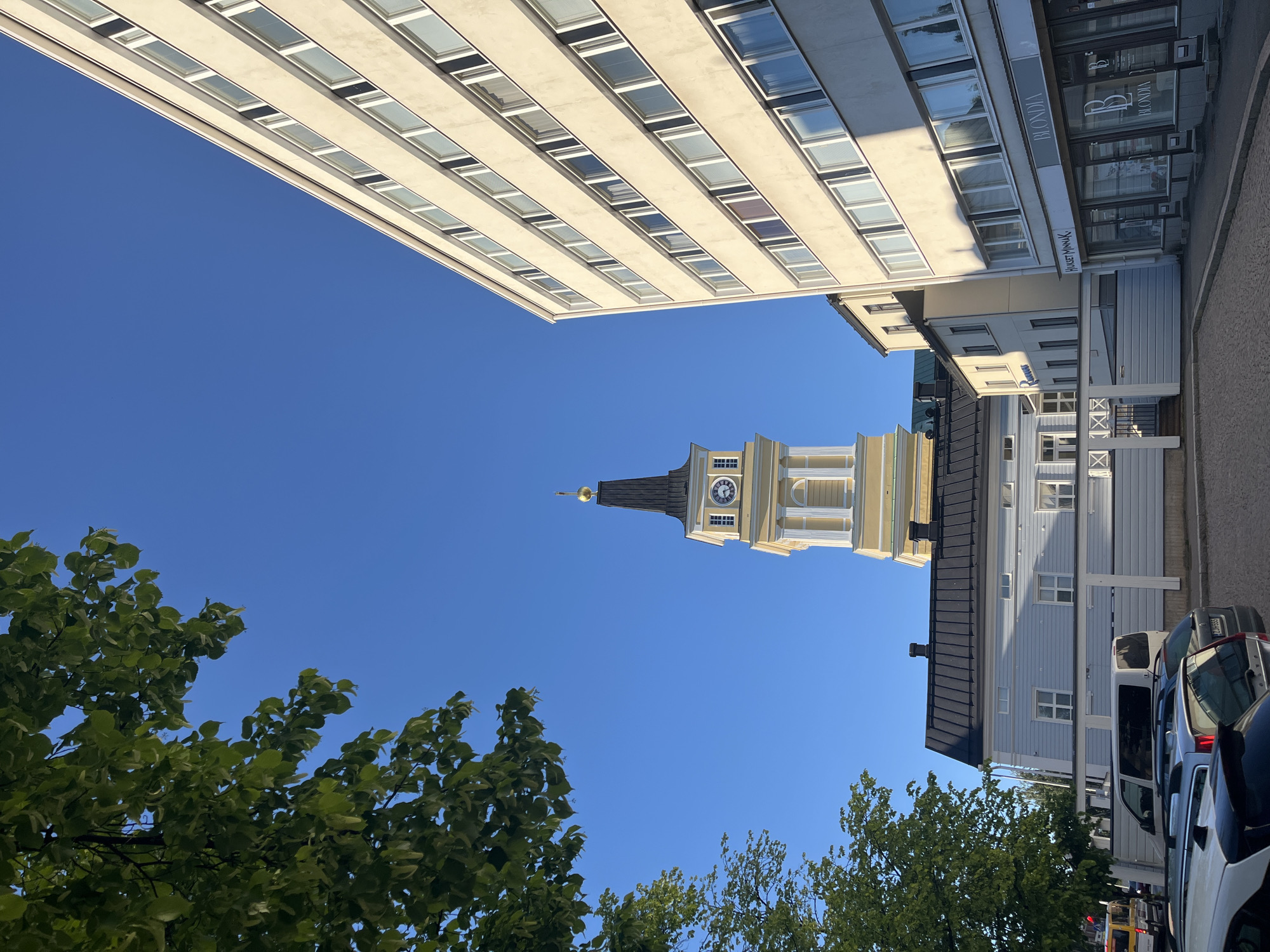 The image captures a street scene in Oulu, Finland, on a clear, bright day. Dominating the right side of the frame is a modern, multi-story building with a light-colored facade and numerous rectangular windows. To the left, a classic yellow and white church tower with a black spire and a clock face stands prominently against the vivid blue sky. In the foreground, a section of a light blue, traditional wooden building with a dark roof is visible, bordered by a white fence. Several cars are parked on a cobblestone street in front of the buildings. Green foliage from a tree hangs into the upper left corner of the image, framing the sky. Shop signs, including "BLONDIA" and "HUUSEN MINNAK," are visible on the ground floor of the modern building. The overall impression is one of an urban environment that blends historical and contemporary architecture.