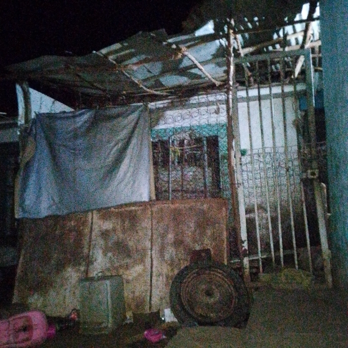 A dilapidated structure with a makeshift roof of corrugated metal and wood. A gray tarp hangs as a partial wall. Faded, rusty walls form the lower part of the building. A metal gate stands to the right of the structure. A large tire sits on the ground in front of the walls. The image is dark, with poor lighting obscuring details of the surrounding area.