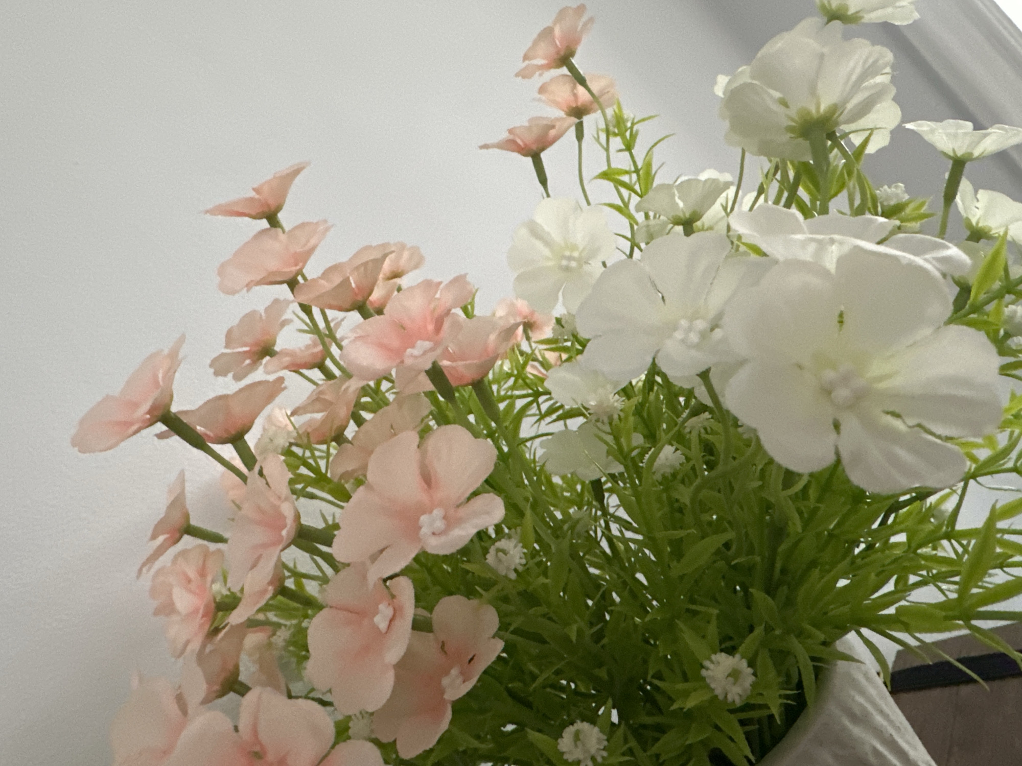 The image displays a close-up view of a floral arrangement, predominantly featuring delicate pink and white artificial flowers. The flowers are clustered together with vibrant green artificial foliage. The arrangement is placed against a plain, light-colored background that appears to be a wall. There is no discernible presence of people or any specific location within Kattakurgan City, Uzbekistan, identifiable from the visual information presented. The focus is solely on the artificial flowers and greenery, creating a simple and decorative composition.